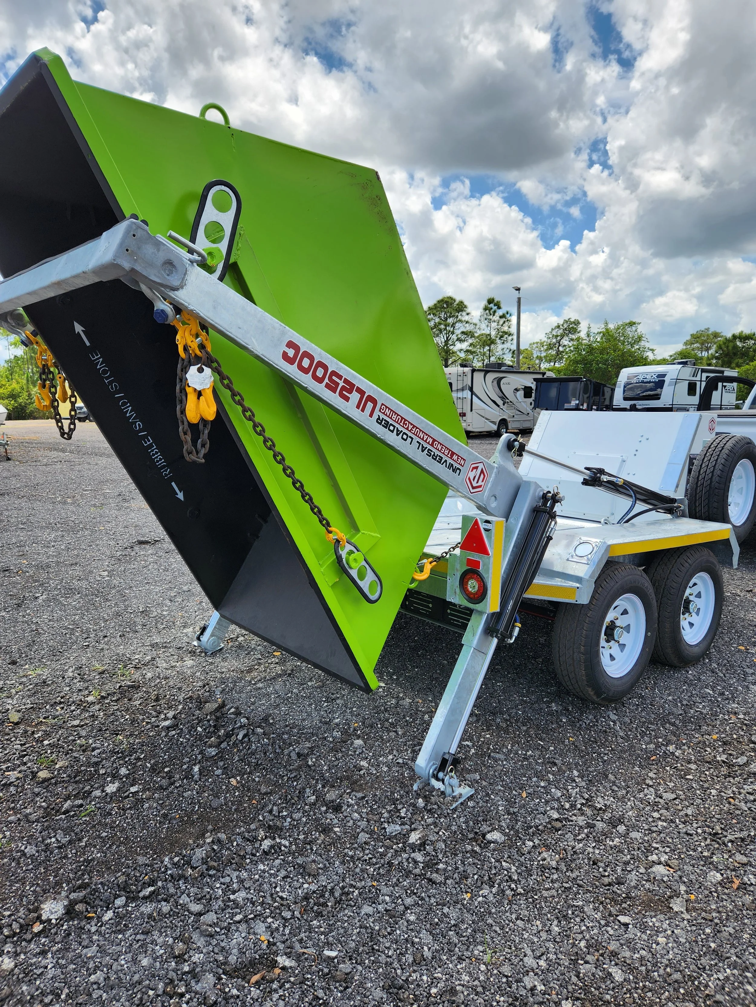 A green and silver mobile portable loader with hydraulic lifting mechanism, parked on gravel ground, with a background of RVs and a partly cloudy sky.