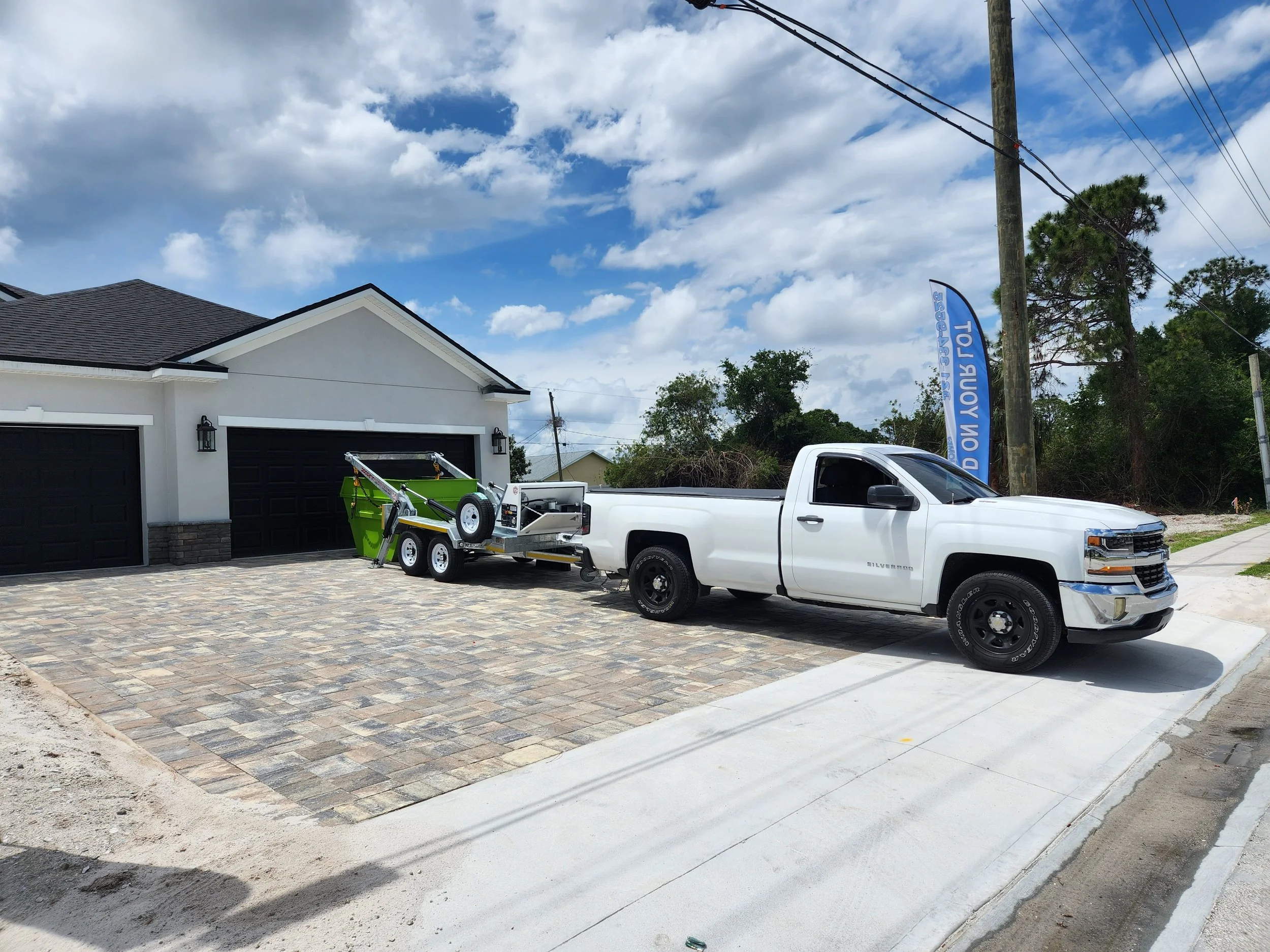 White pickup truck parked on a driveway next to a garage, towing a green trash compactor with a trailer, under a partly cloudy sky in a residential neighborhood.