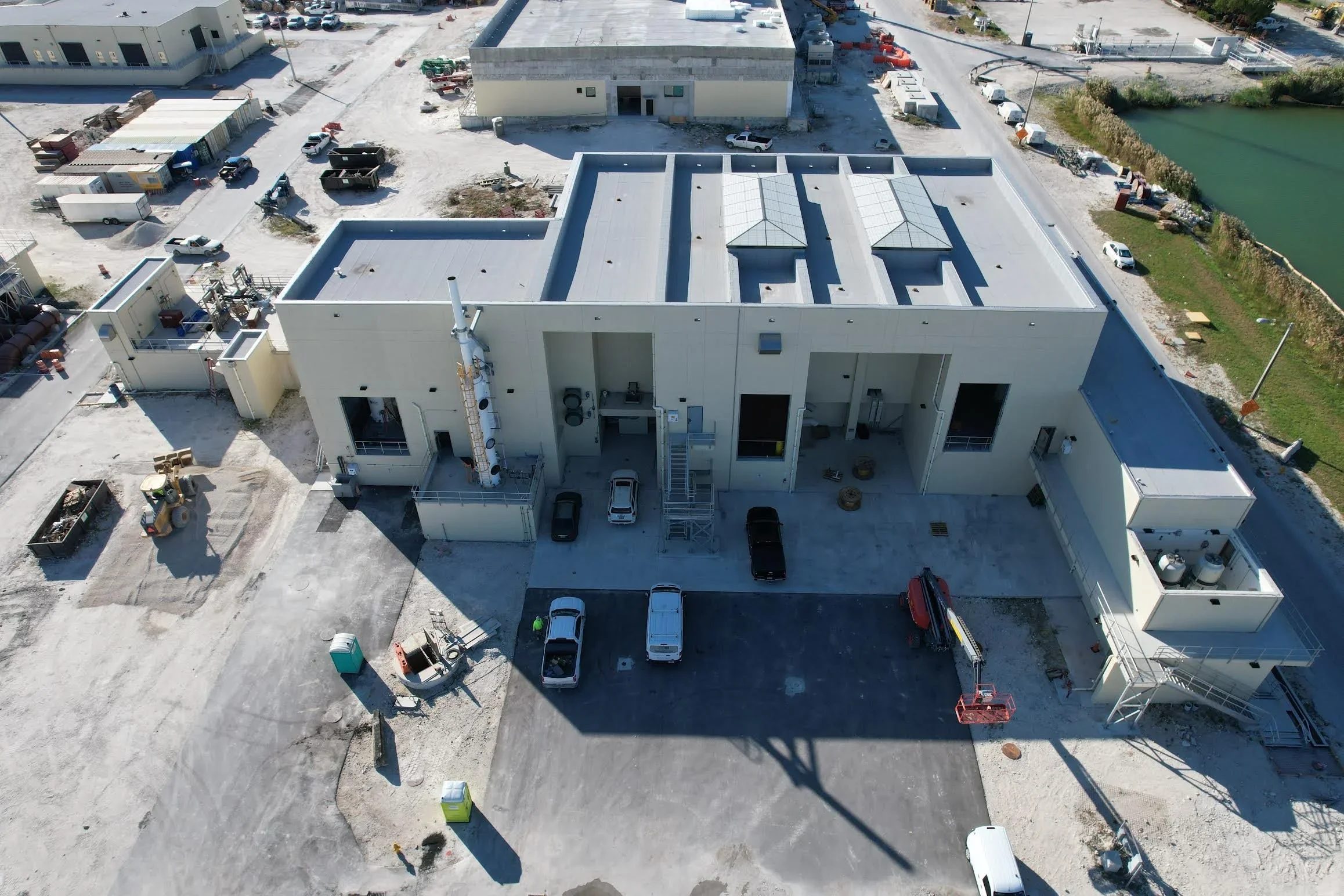 An aerial view of a construction site with a large white building in progress, several parked vehicles, construction equipment, and gravel areas, with a water body on the right side.