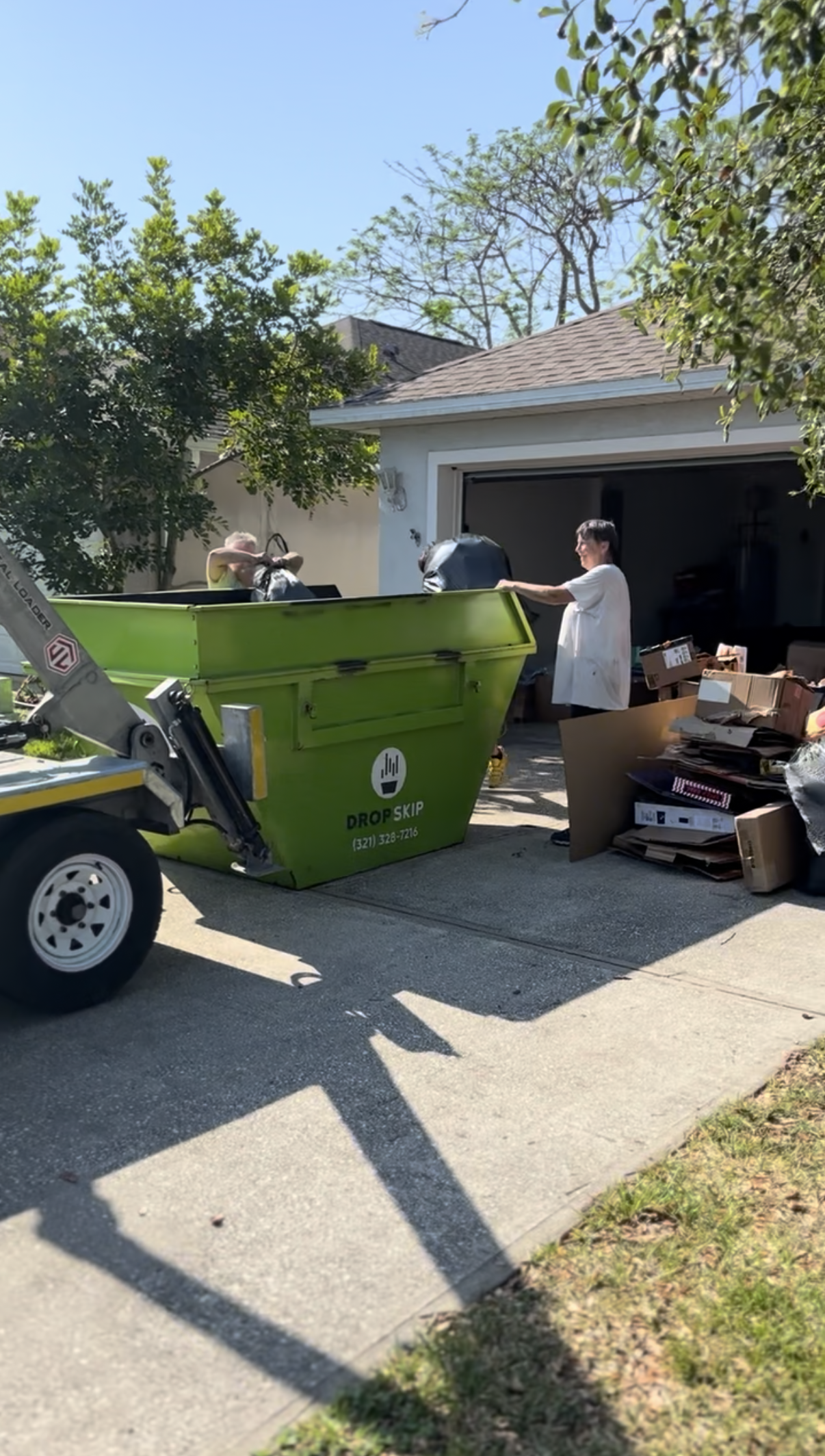 A person is throwing trash into a large green dumpster in a driveway, with another person inside the dumpster. There are boxes and trash bags piled beside the dumpster, and a truck is nearby.