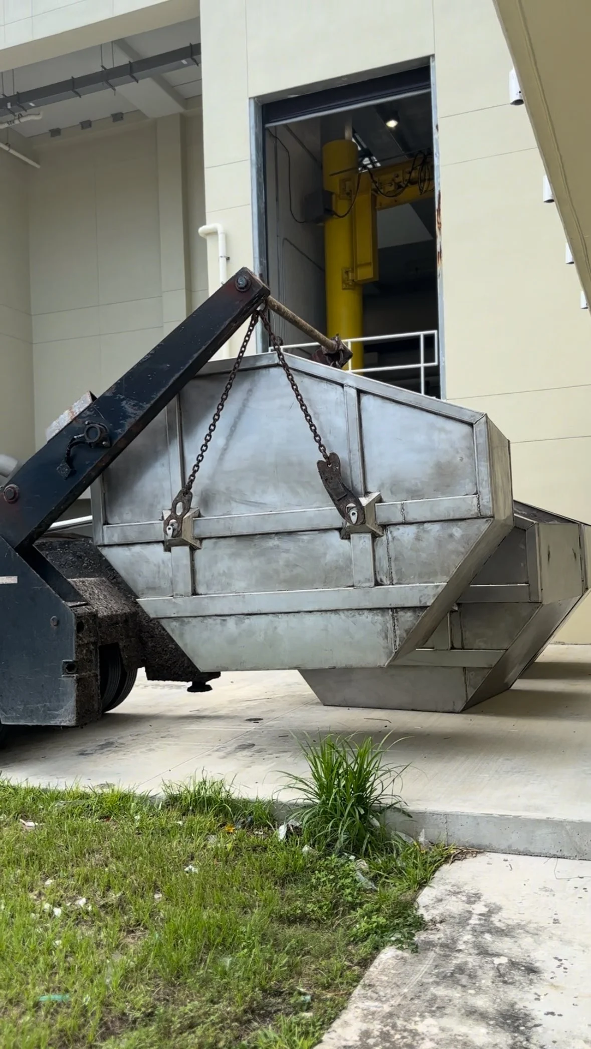 Heavy-duty metal dumpster being lifted by a construction lift outside a building with an open door and yellow equipment inside.