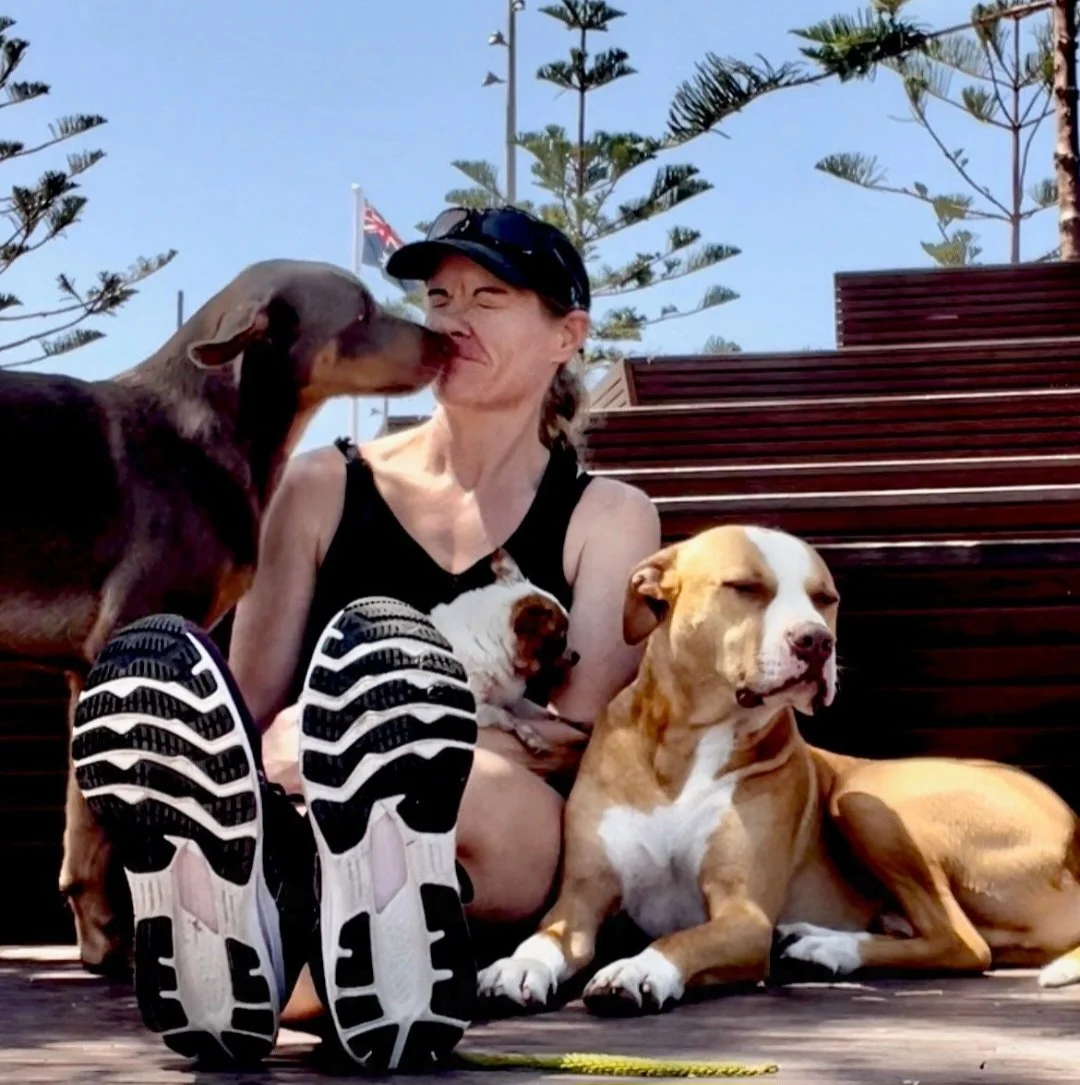 A woman sitting on wooden steps outdoors, surrounded by four dogs. One dog is licking her face, another is resting against her, and the other two are lying nearby. She is smiling with her eyes closed, wearing a black tank top, black cap, and running shoes. The background shows trees, a flag, and a clear blue sky.