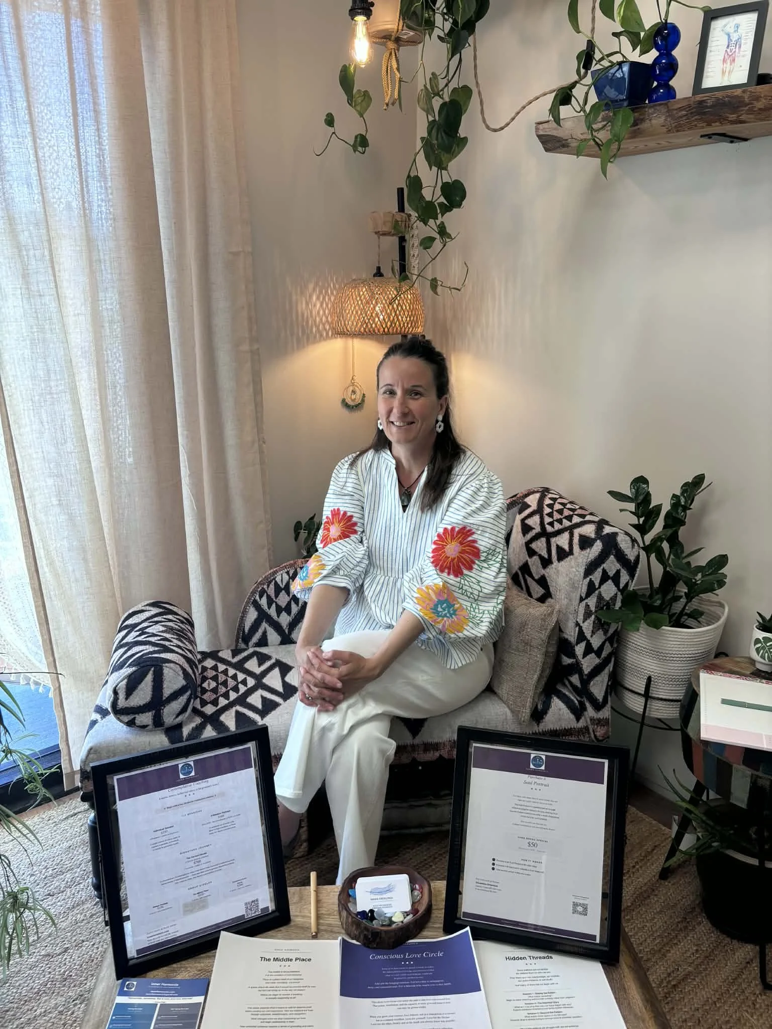 A woman with dark hair and earrings, wearing a striped shirt with floral embroidery, sits on a patterned sofa in a room decorated with plants, framed pictures, and framed informational sheets on some tables in front of her.