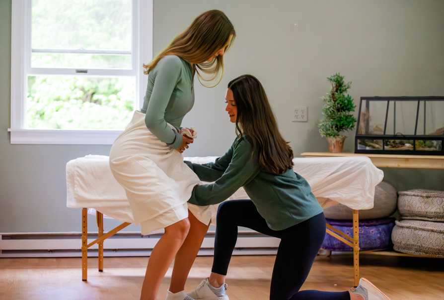 Two women in a massage or therapy room, one sitting on a massage table, the other kneeling beside her, holding her hand, with a window, plants, and furniture in the background.