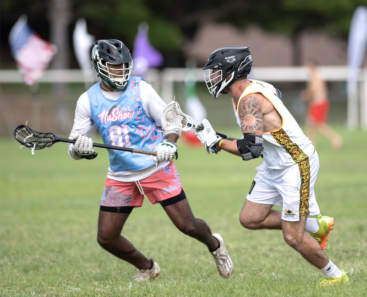 Two lacrosse players wearing helmets and protective gear, competing for ball possession on a grassy field during a game. One player is in a light blue and pink uniform, the other in a white and yellow uniform, with flags and spectators blurred in the background.