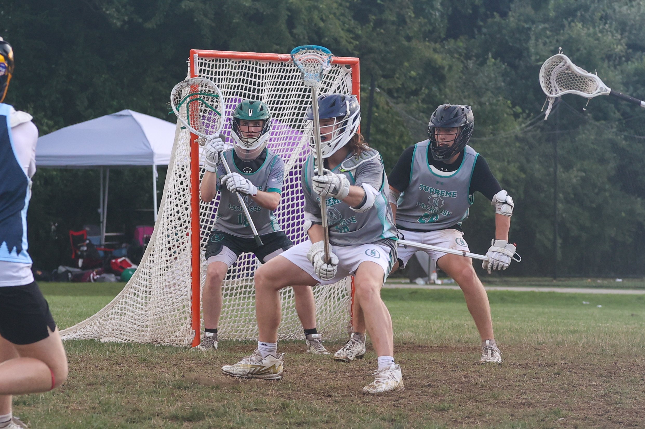 Lacrosse players in gray jerseys forming a defensive wall in front of the goal, with one player holding a lacrosse stick and other players preparing to defend during a game on grassy field.
