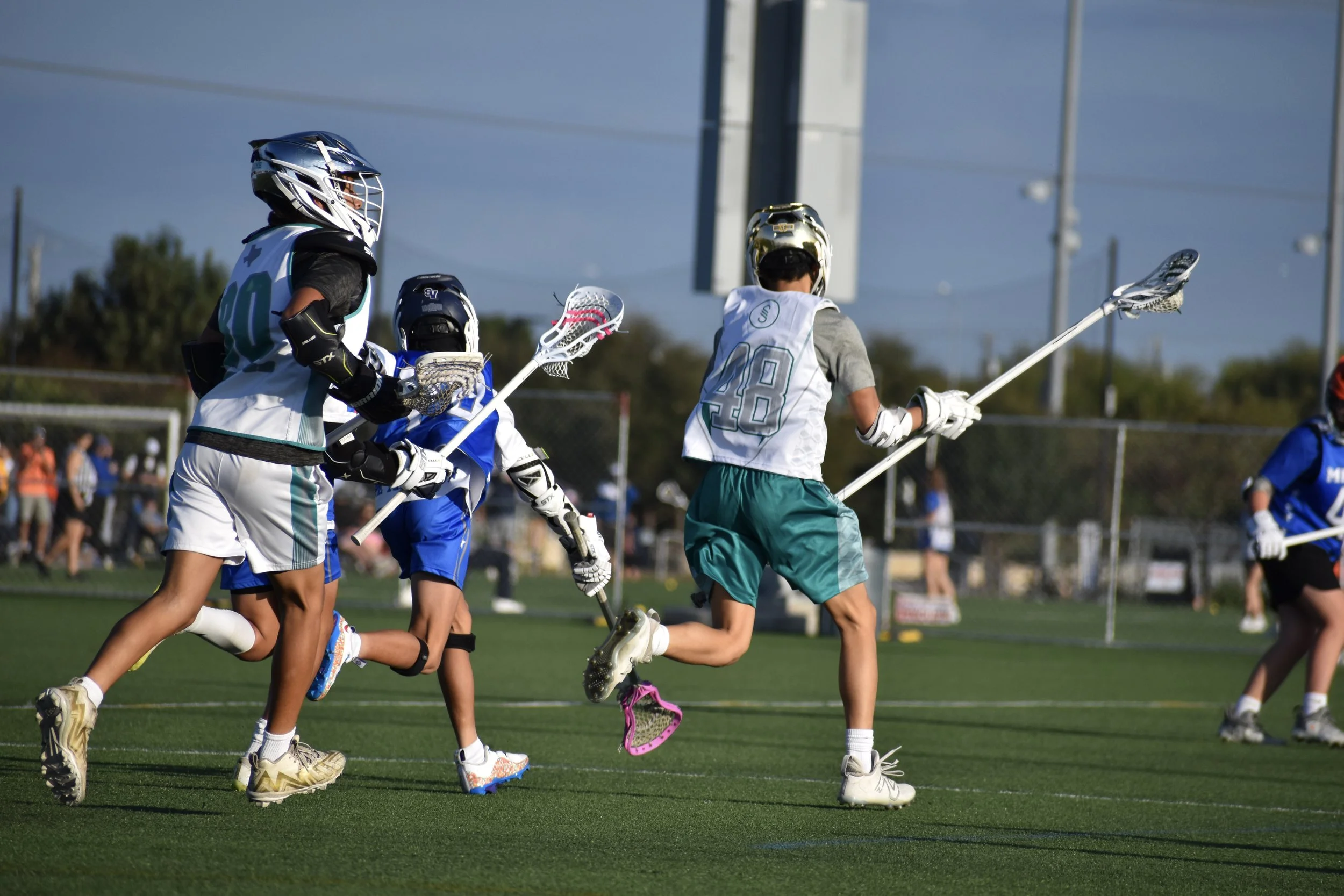 Kids playing lacrosse on a field with a chain-link fence and spectators in the background.