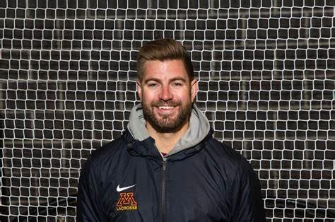 A man smiling, wearing a dark jacket with a 'Minnesota lacrosse' logo, standing in front of a black net background.