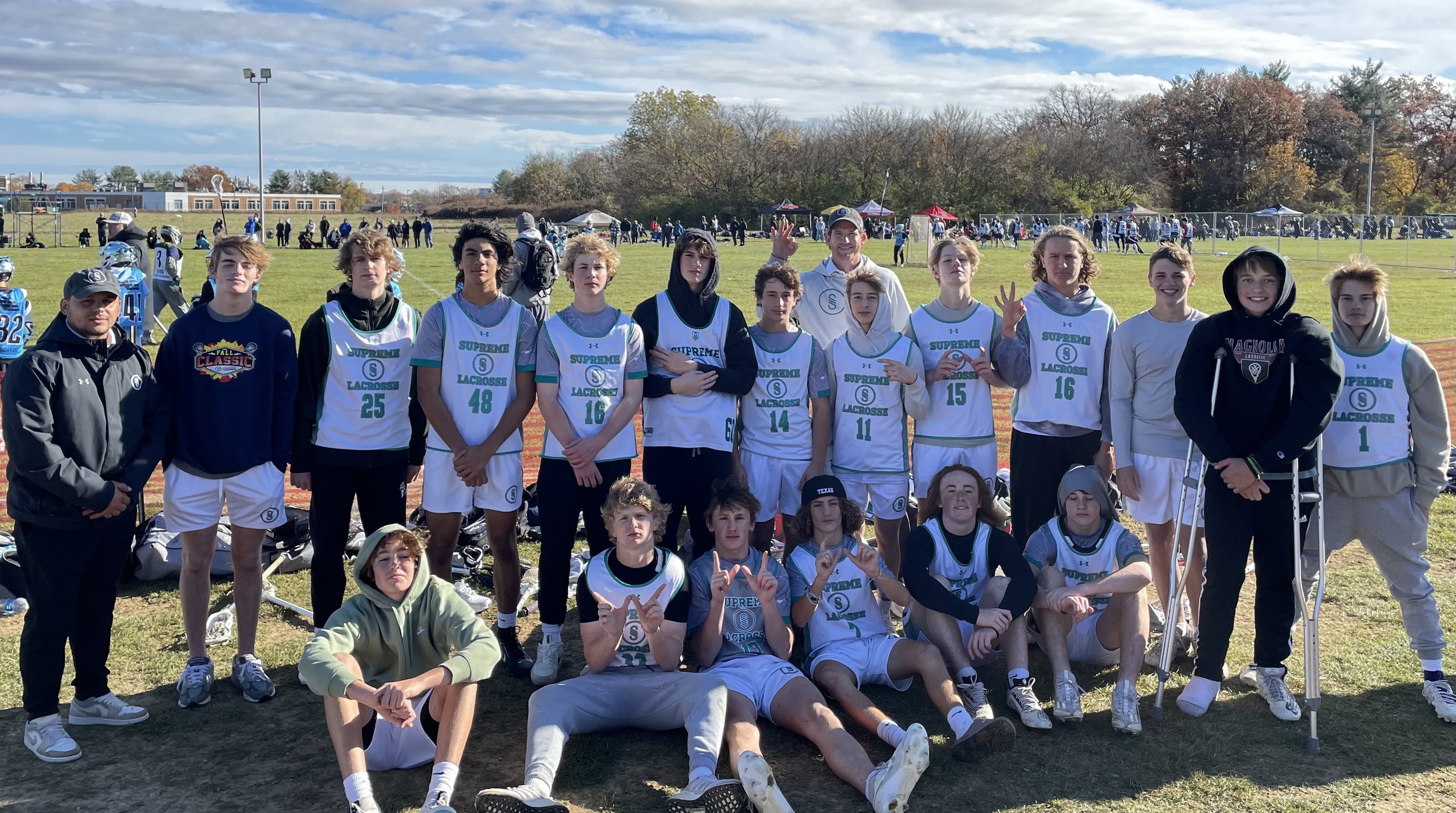 Youth lacrosse team and coaches posing for a group photo outdoors on a sports field with trees and a cloudy sky in the background.