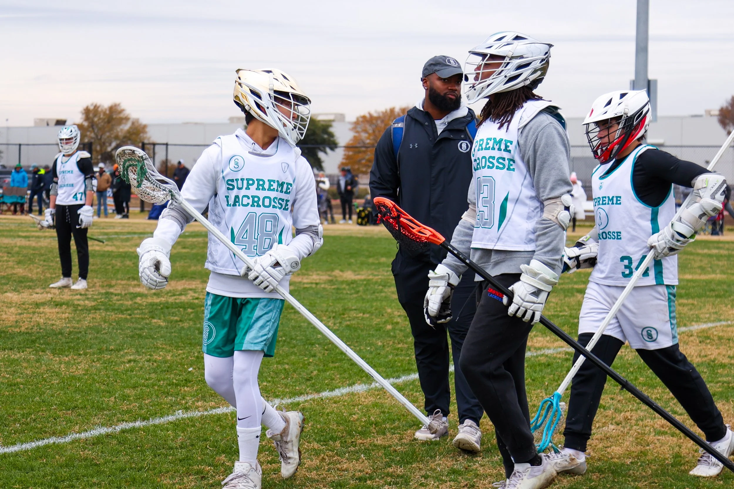 Lacrosse players and coach on a field during a game, wearing white jerseys with green lettering that read 'Supreme Lacrosse.'