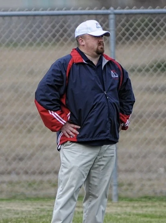 A man standing outdoors in front of a chain-link fence, wearing a navy and red jacket, beige pants, and a white cap.