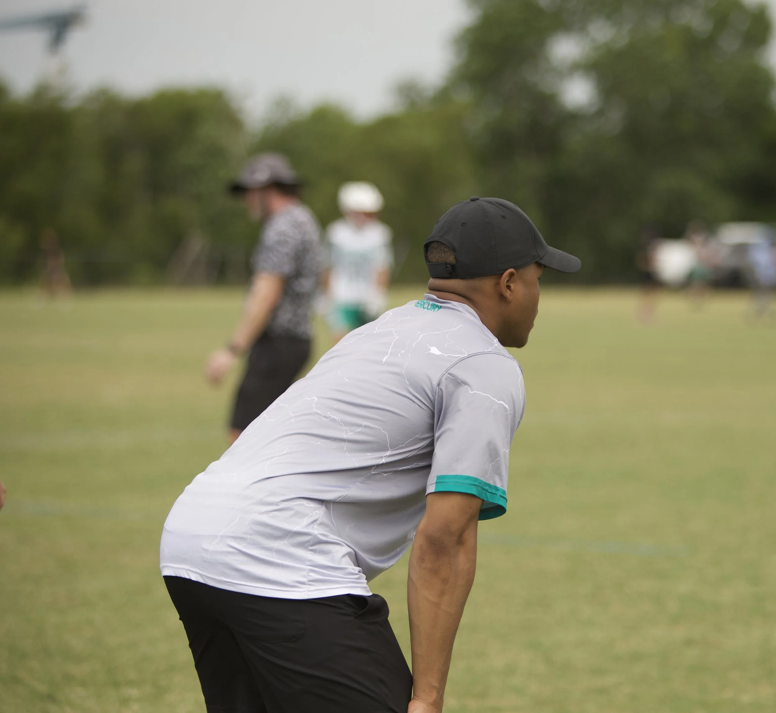 Young man in a white and teal t-shirt and black shorts bending forward on a grassy field during an outdoor event.