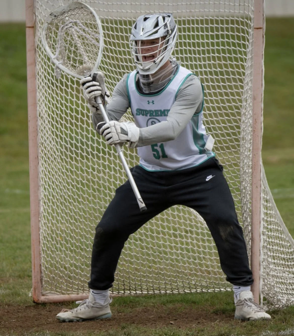 Lacrosse goalie standing in front of a lacrosse goal net, holding a lacrosse stick, wearing protective helmet, gloves, and sports attire.
