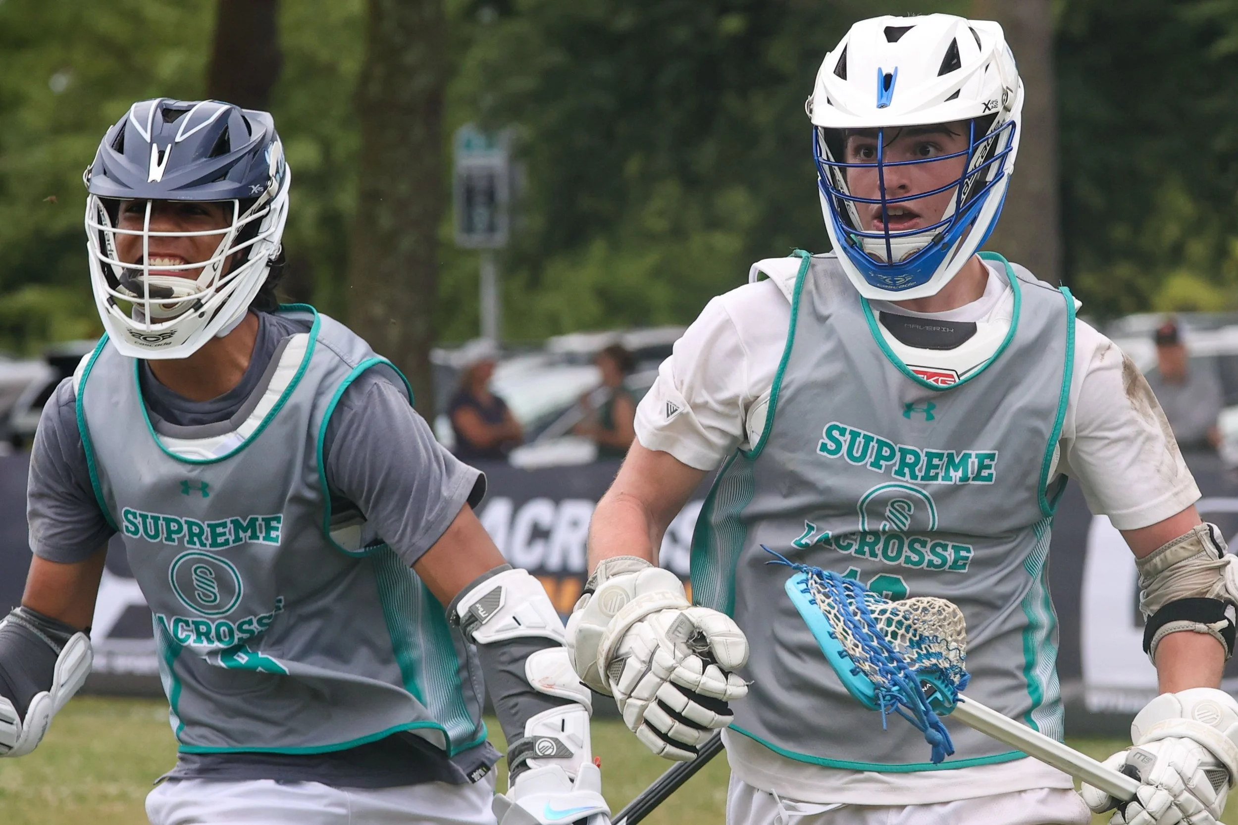 Two male lacrosse players, wearing helmets and jerseys with 'Supreme Lacrosse' on the front, are on a field with trees in the background. They are holding lacrosse sticks, and appear to be engaged in a game.