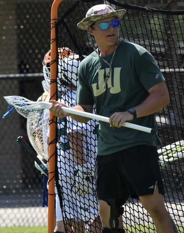 A man holding a lacrosse stick standing next to a lacrosse goal during a practice or game, wearing sunglasses, a camouflage hat, a green shirt, and black shorts.