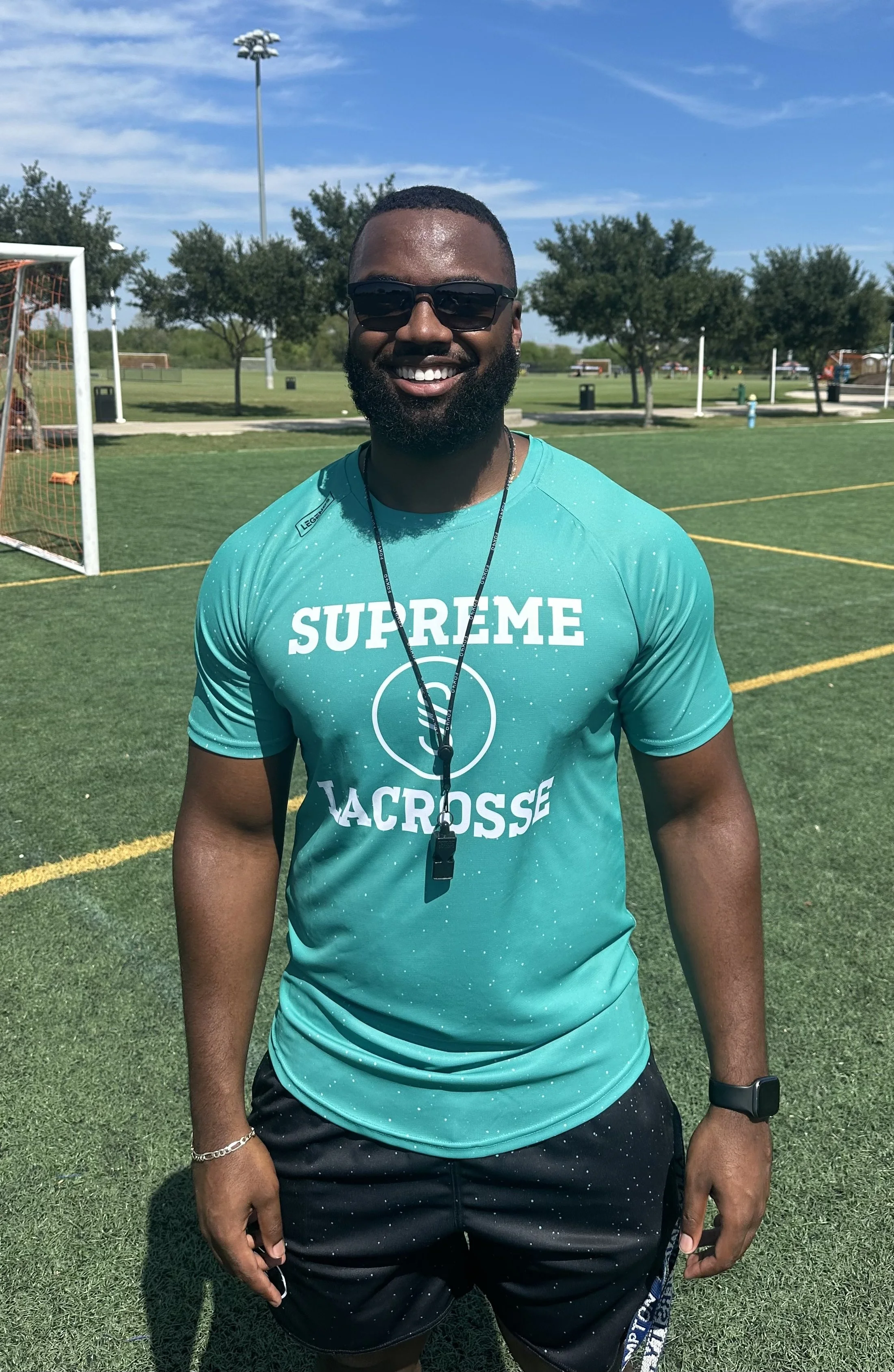 Young man wearing sunglasses, a turquoise Supreme Lacrosse t-shirt, black shorts, and a whistle around his neck, standing on a sports field with goalpost and trees in the background on a sunny day.