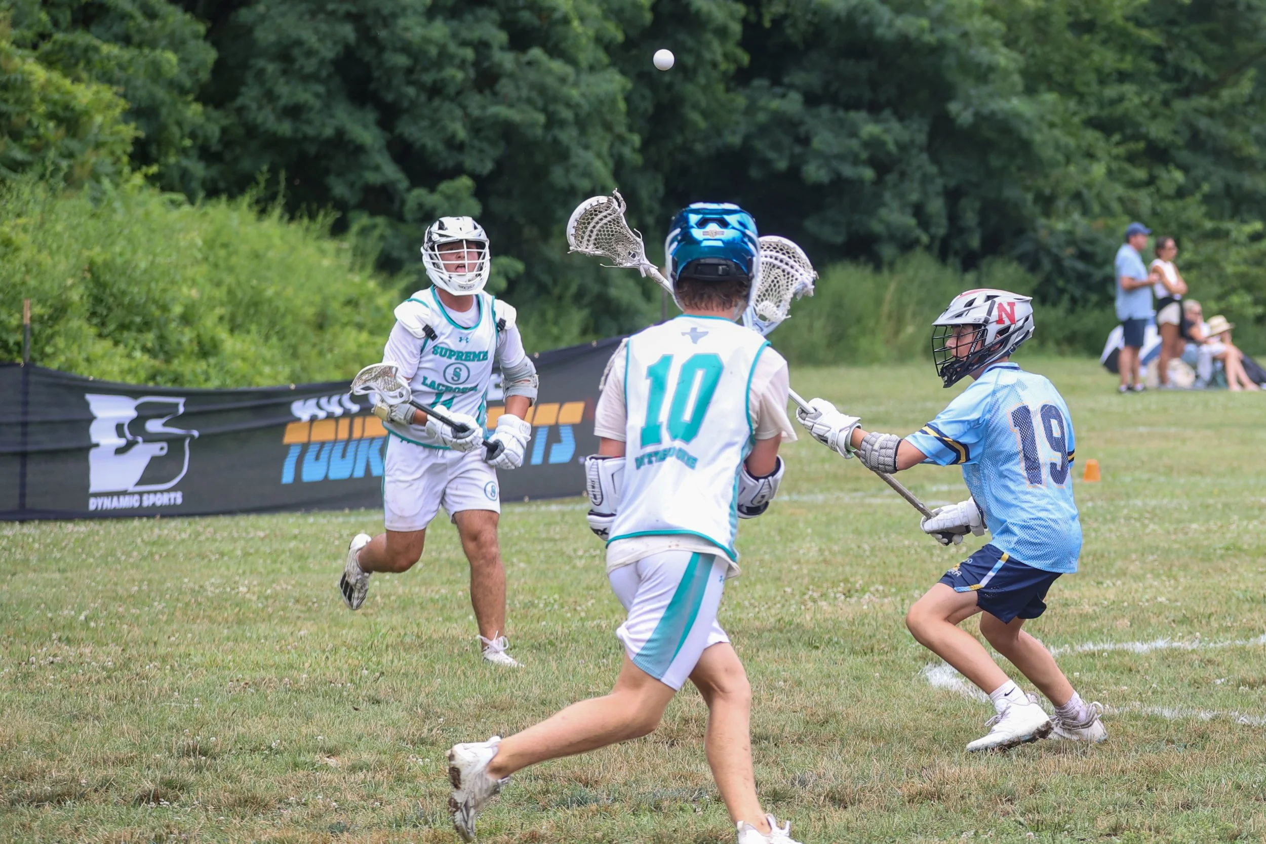 Young boys playing lacrosse outdoors, wearing helmets and sports uniforms, with a lacrosse ball in midair.