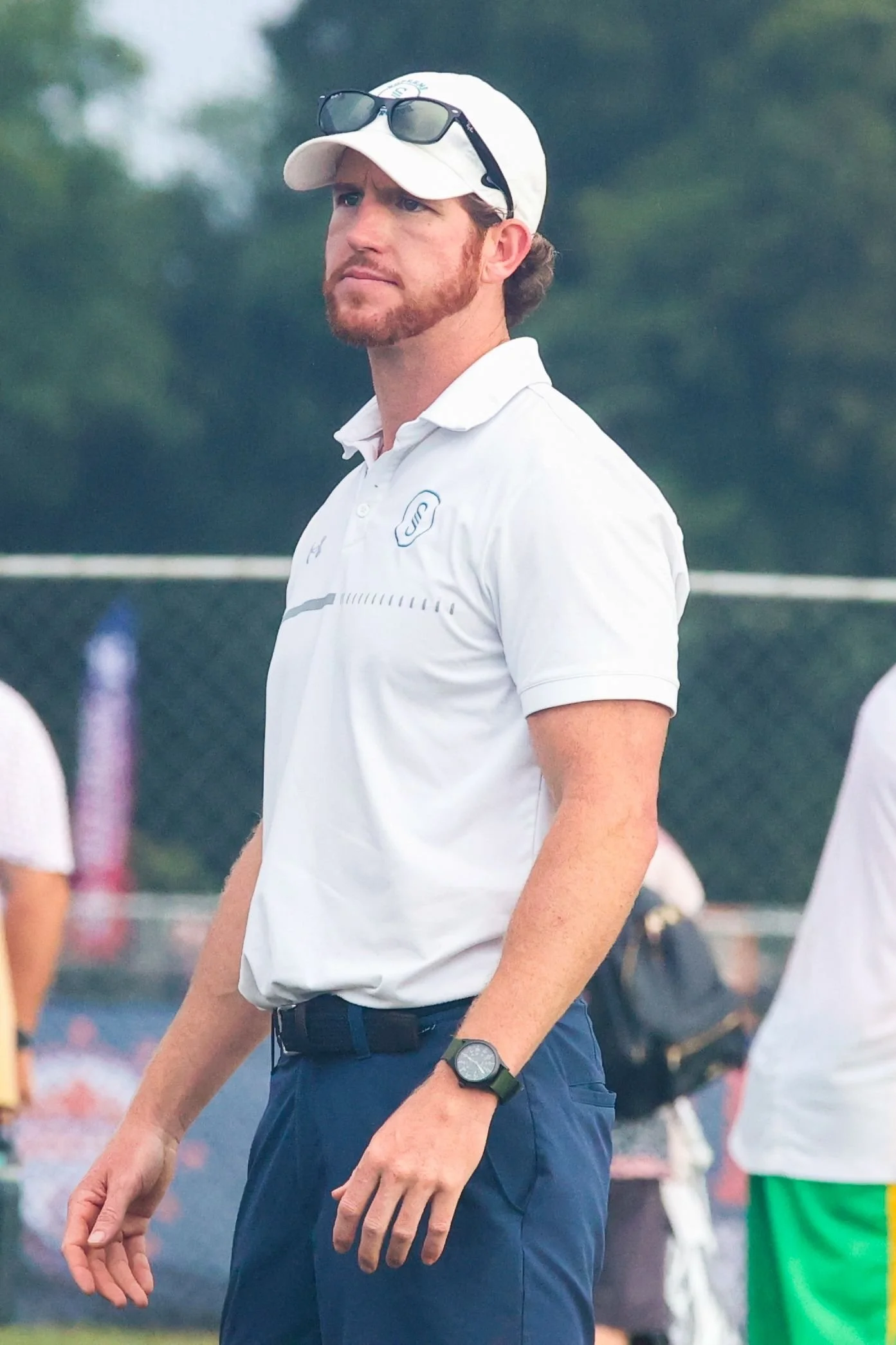 A man wearing a white polo shirt, baseball cap, sunglasses on the cap, and a wristwatch, standing outdoors on what appears to be a sports field.