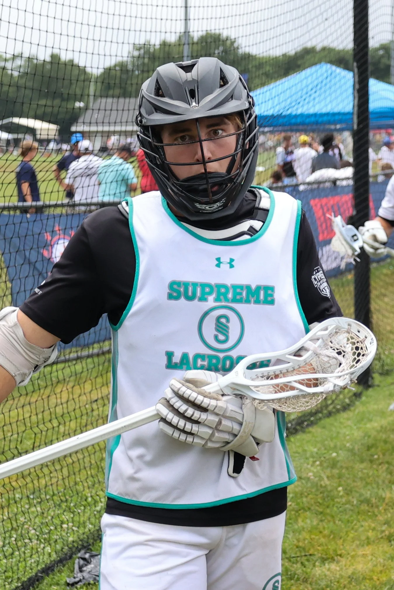 A lacrosse player wearing a black helmet and a white jersey with teal accents, standing on a field with a net fence and other people in the background.