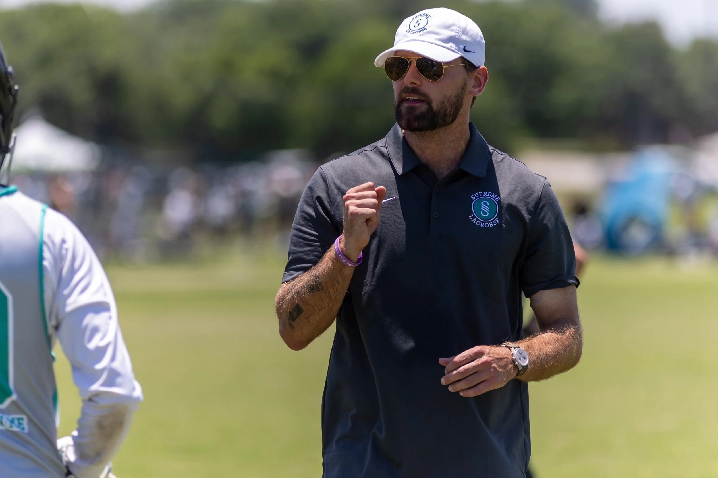 A man wearing sunglasses, a white cap, a wristwatch, and a dark polo shirt with a lacrosse logo is standing outdoors on a grassy field. He appears to be coaching or giving instructions.