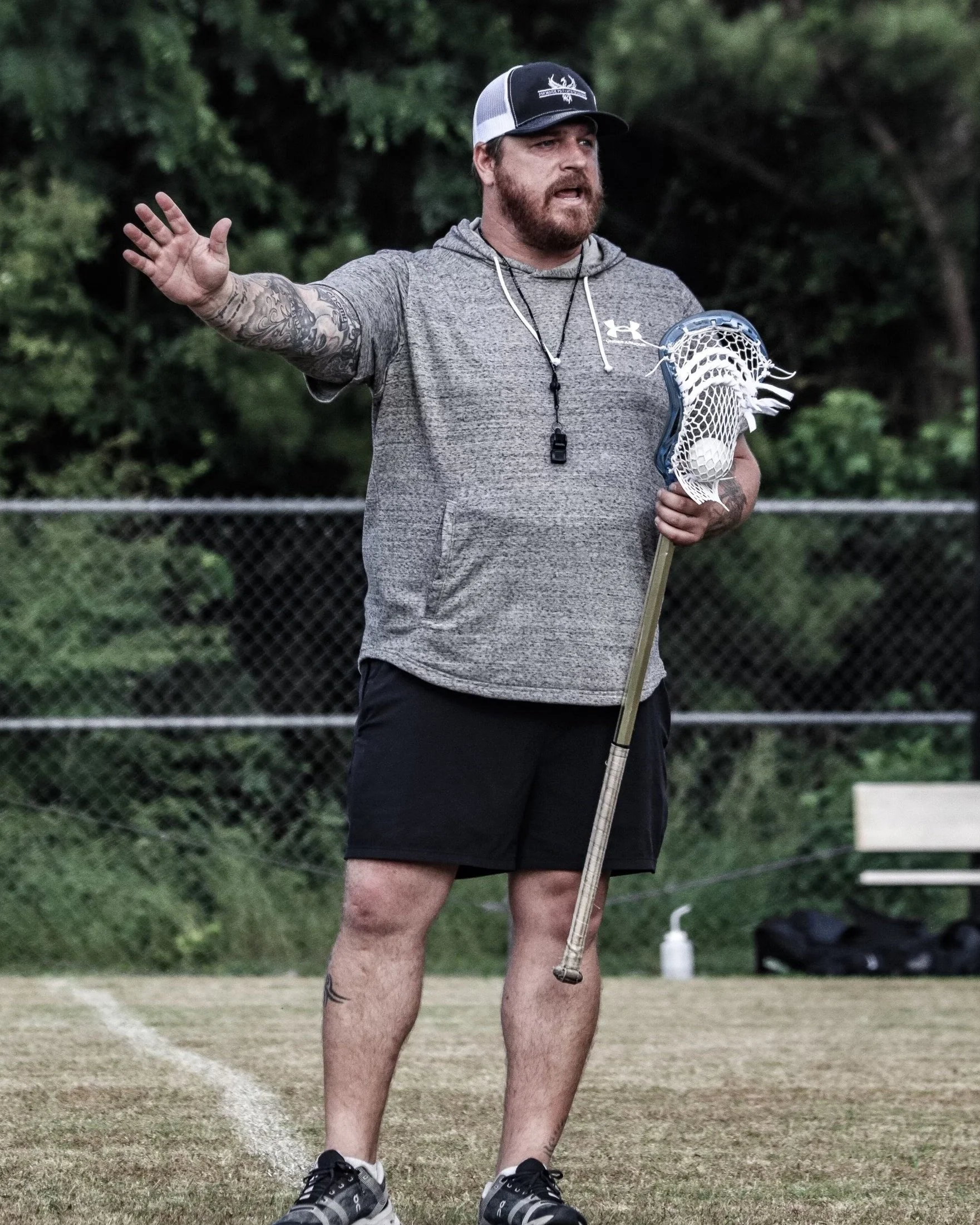 A man with a beard and tattoos, wearing a gray hoodie, black shorts, and a black and white cap, holding a lacrosse stick and ball, standing on a sports field, gesturing with his left hand.