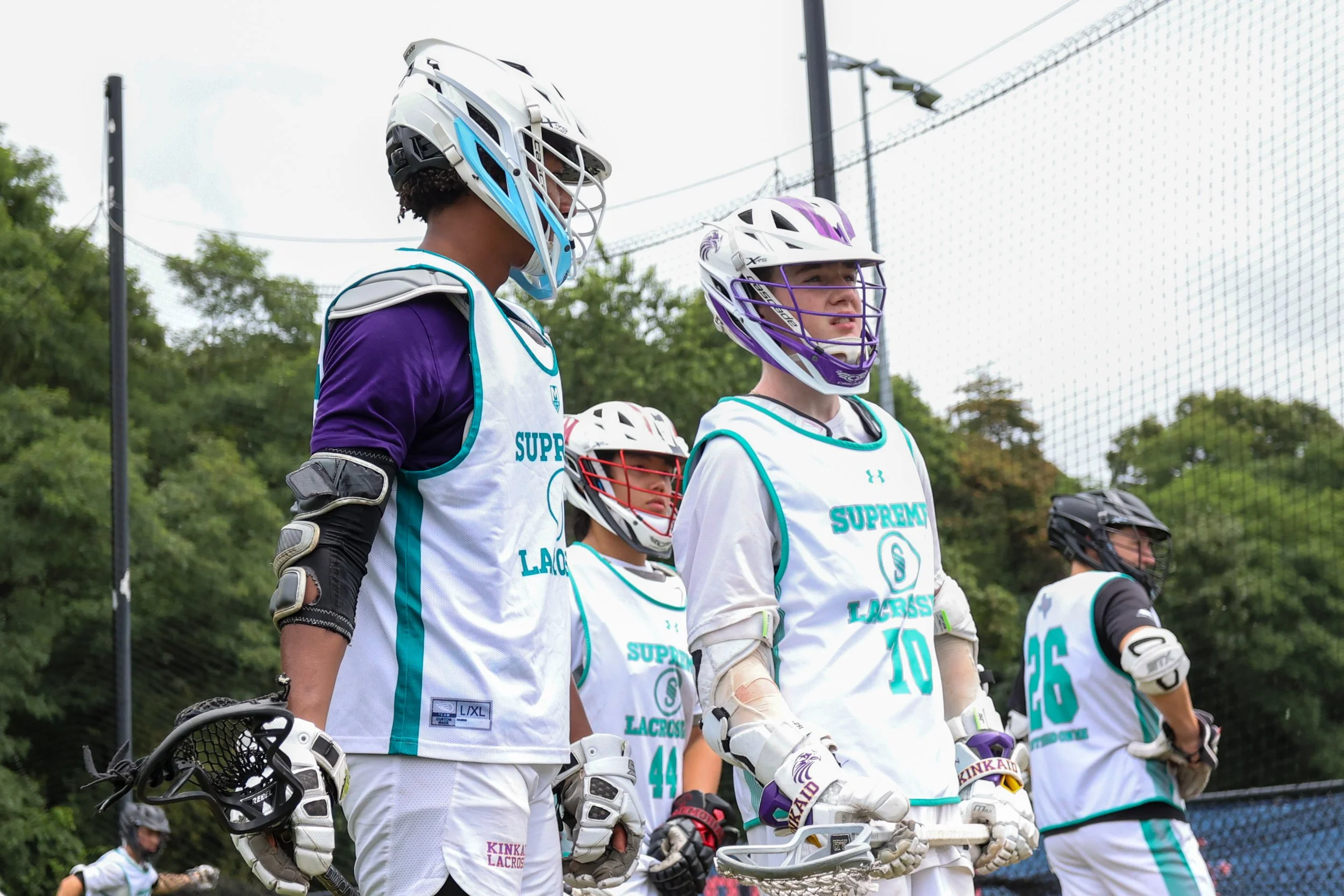 Four youth lacrosse players in white and teal jerseys with helmets, standing on a lacrosse field.