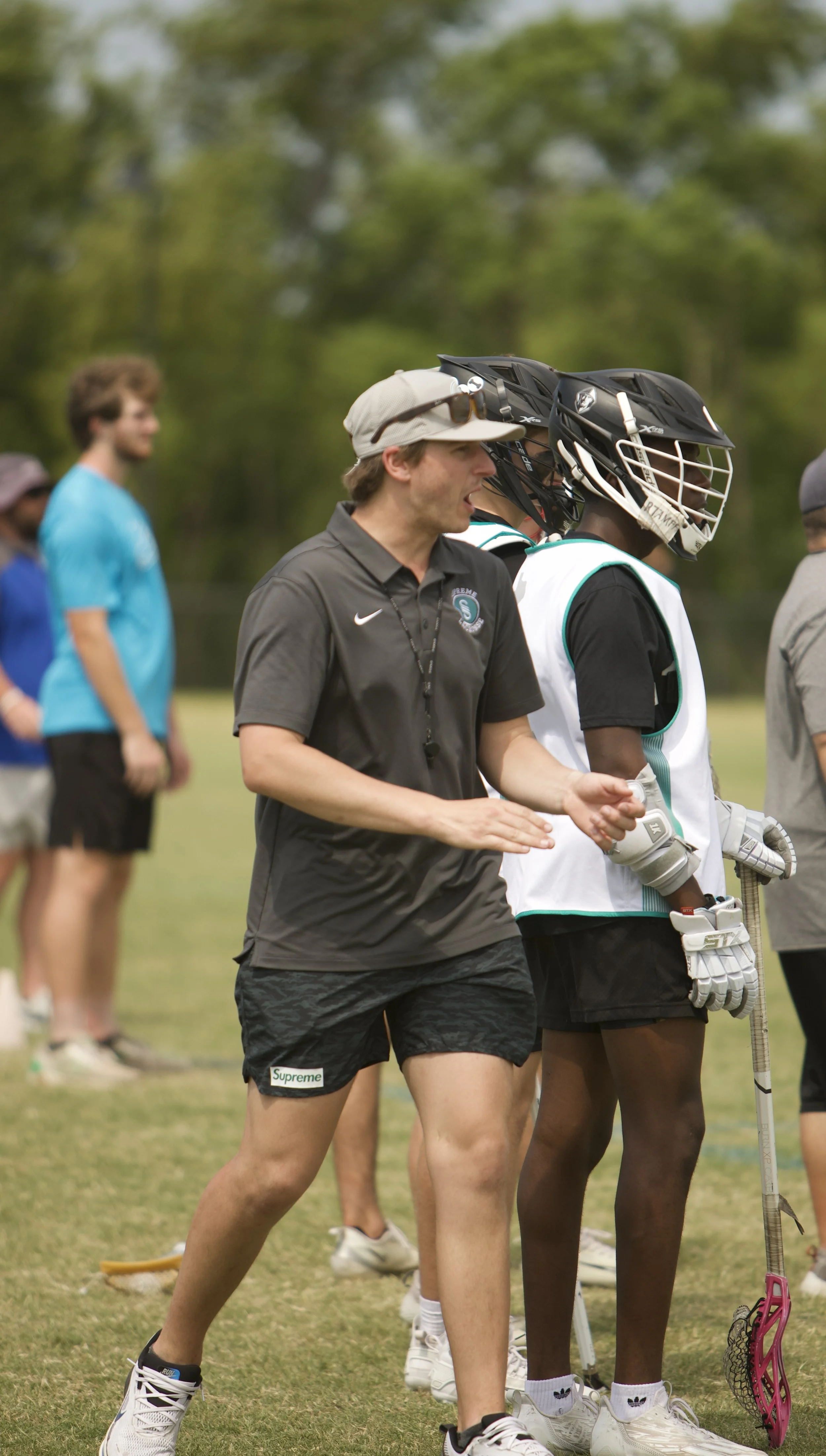 Lacrosse coach giving instructions to players on a grassy field, with other spectators in the background.