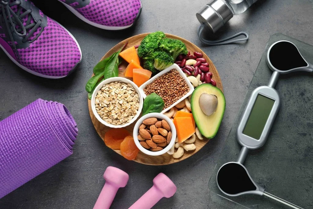 Healthy lifestyle concept with a wooden plate of nutritious food including oats, almonds, avocado, broccoli, beans, and more surrounded by exercise equipment like pink sneakers, a purple yoga mat, pink dumbbells, a water bottle, and a digital scale on a dark surface.