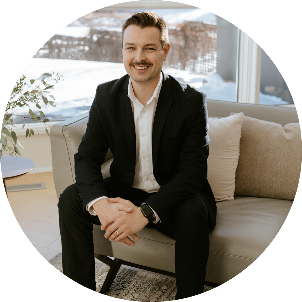 Best Financial Advisor Winnipeg Kevin Rawluk sitting relaxed on a tan sofa smiling. He is wearing a black suit with no tie.