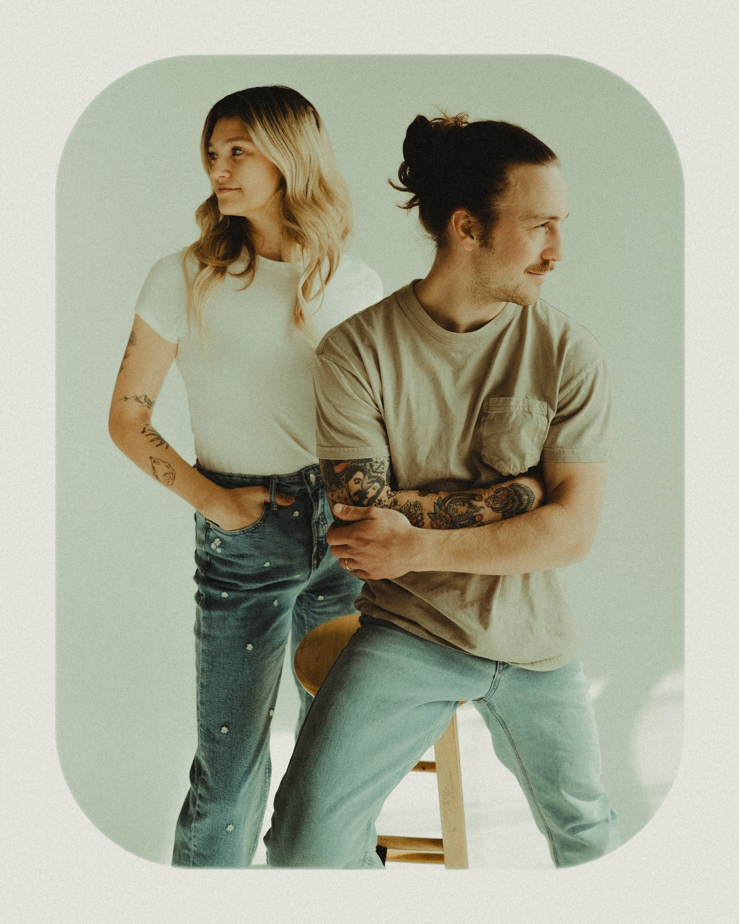 A man and woman with tattoos pose against a light green background. The woman stands with her hands in her pockets, looking to the side. The man sits on a stool, arms crossed, also looking to the side.