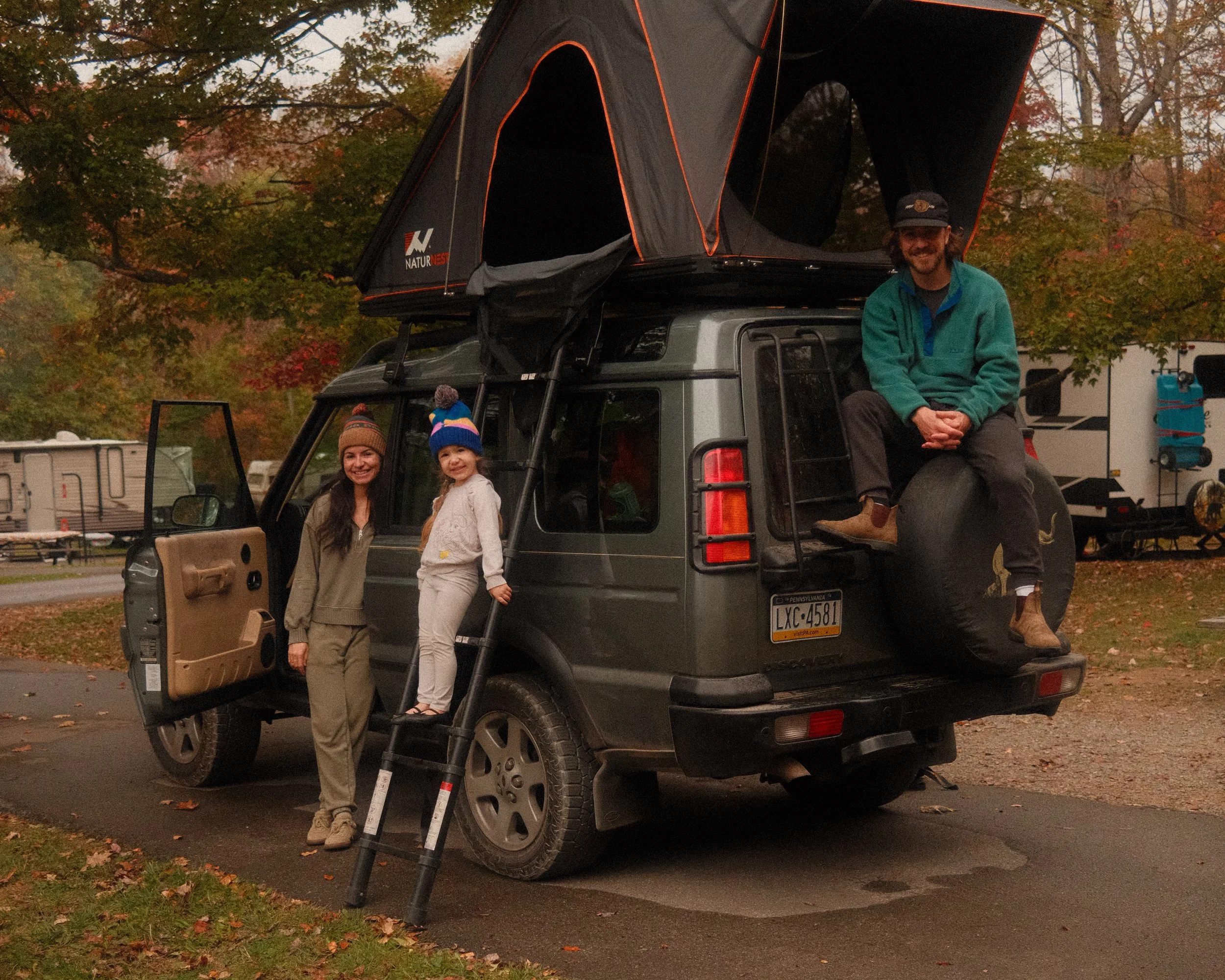 A family of three enjoying fall camping. They are building a JavaScript log cabin next to their car. The scene is set in an autumn park with colorful leaves, a camper, and a trailer in the background.
