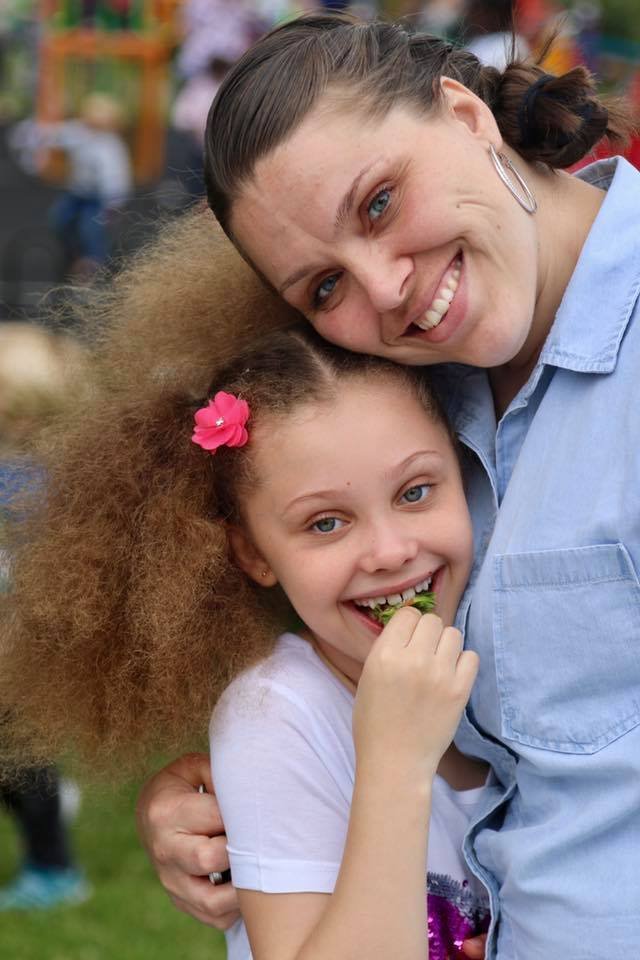 A woman and a young girl smiling outdoors. The girl has a pink flower in her hair and is holding a green object near her mouth. The woman hugs her from the side, both appearing happy.