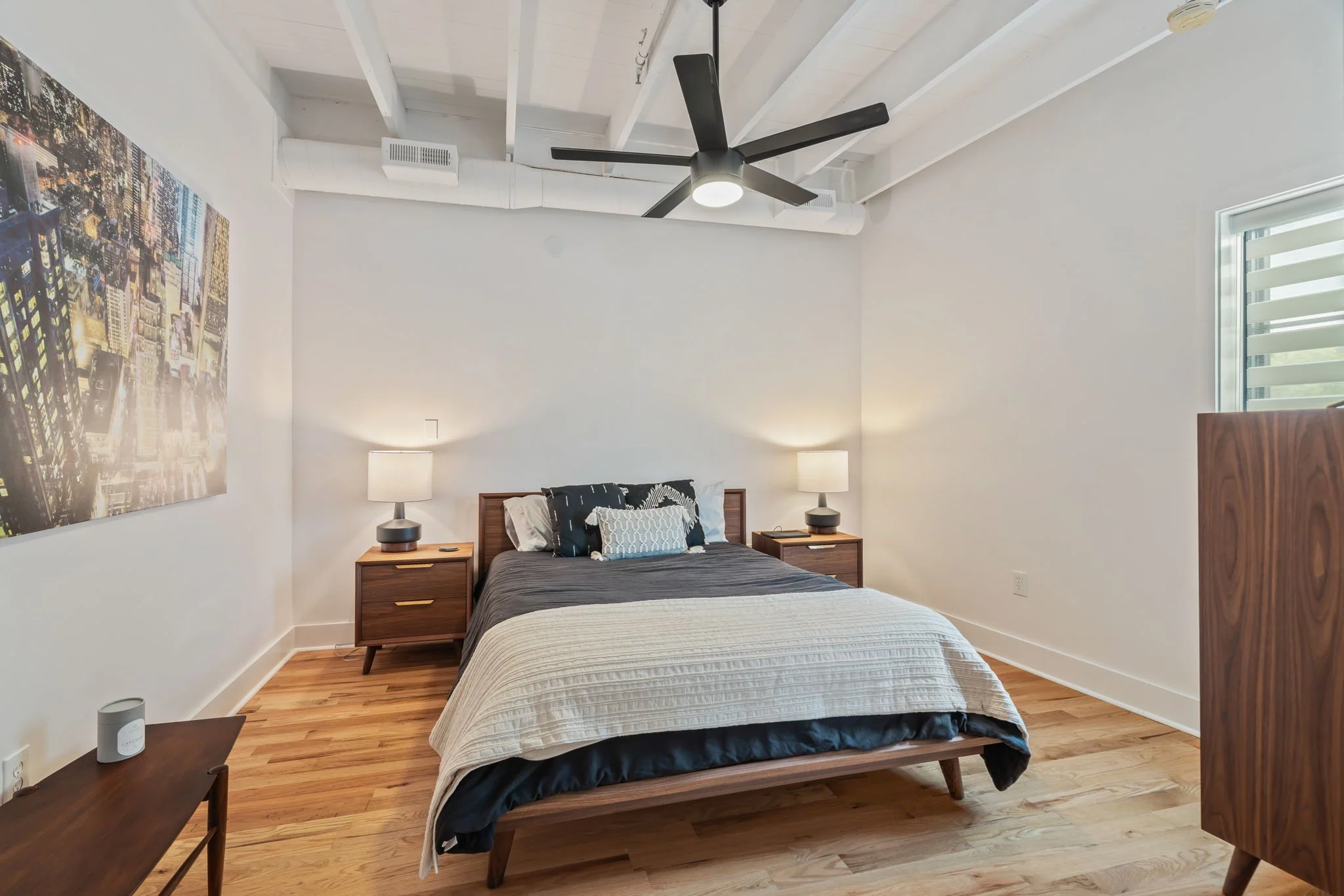Modern bedroom with a wooden bed frame, dark gray bedding, and multiple pillows, flanked by wooden nightstands with lamps, a ceiling fan, and a cityscape artwork on the wall.