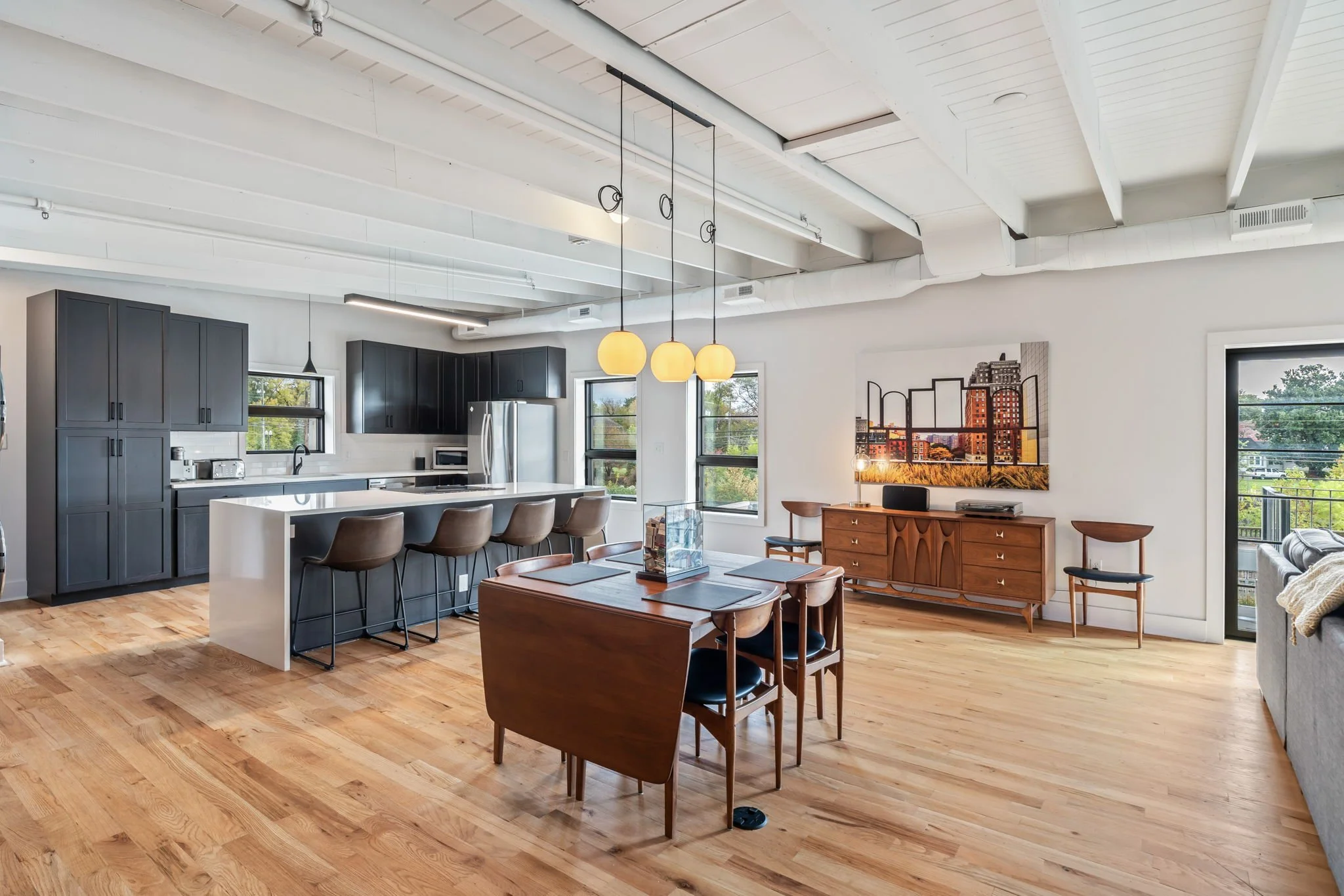 Open-concept living space with a kitchen, dining area, and seating. The kitchen has dark cabinets, white countertops, a refrigerator, and barstools. The dining area features a wooden table with chairs. There's a sideboard with a large framed cityscap