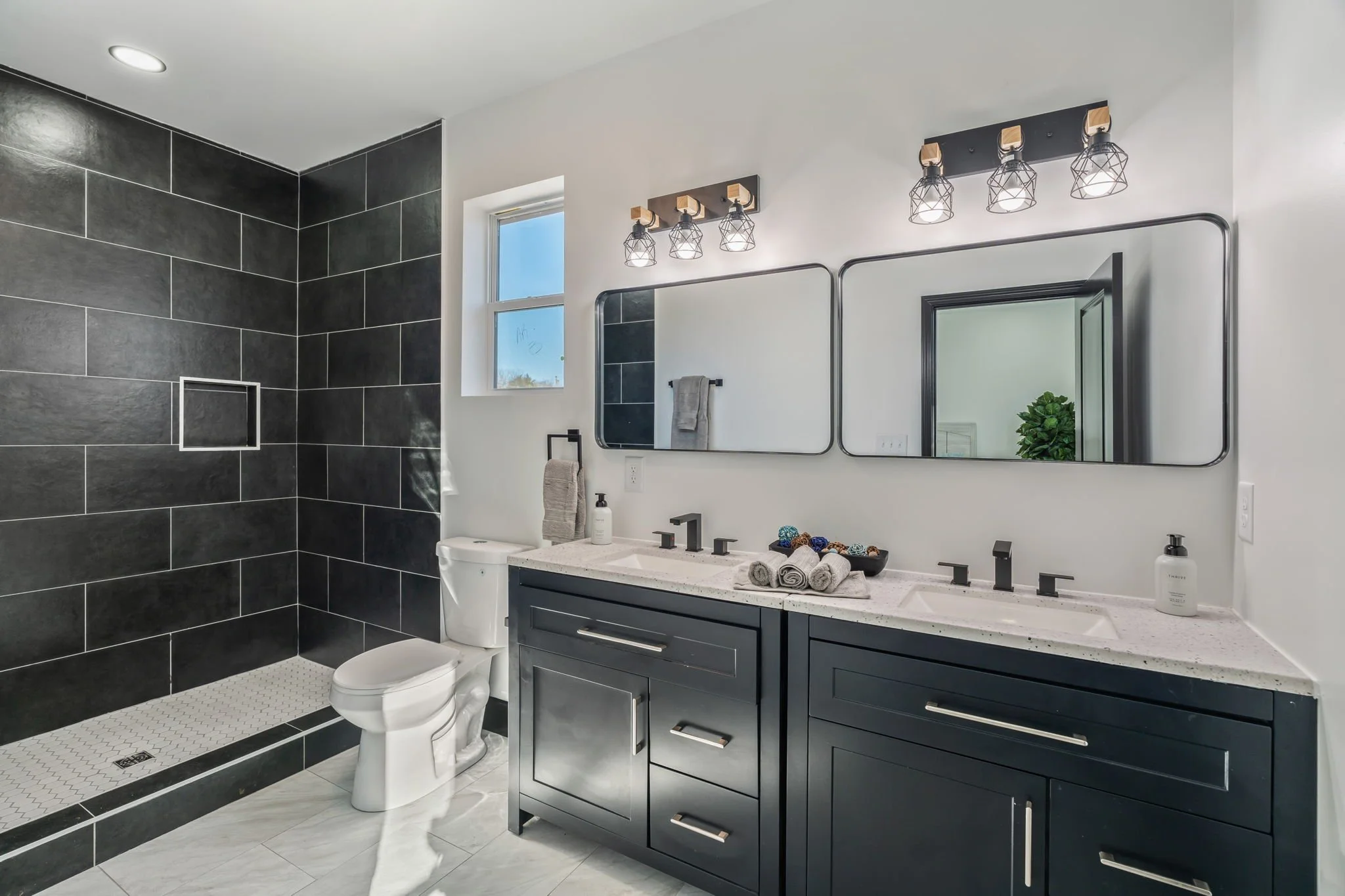 Modern bathroom with double vanity, black cabinets, large mirrors, two black sinks, black faucets, decorative towels, and a black tiled walk-in shower with gray grout and a niche, white hexagonal floor tiles, white walls, small window, and modern lig