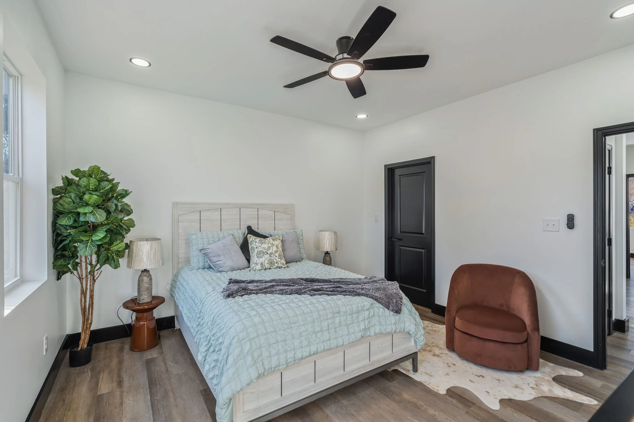 A bedroom with a white bed, green pillow, gray pillows, a gray blanket, and two bedside lamps on bedside tables. There's a tall green plant with large leaves in the corner and a brown round chair near the door. The room has white walls, black door, a