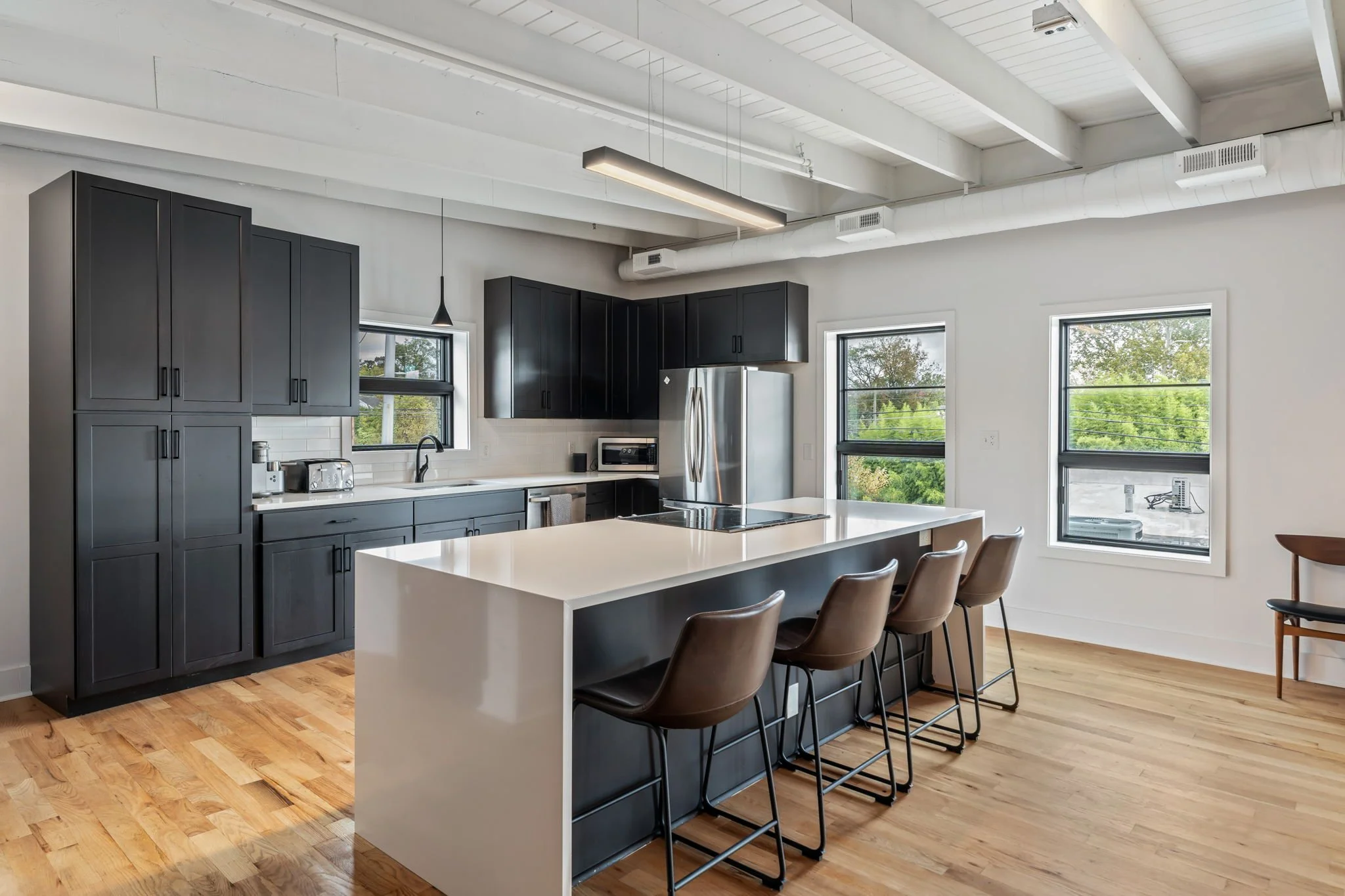 Modern kitchen with black cabinets, white island, wood flooring, stainless steel appliances, and large windows showing greenery outside.