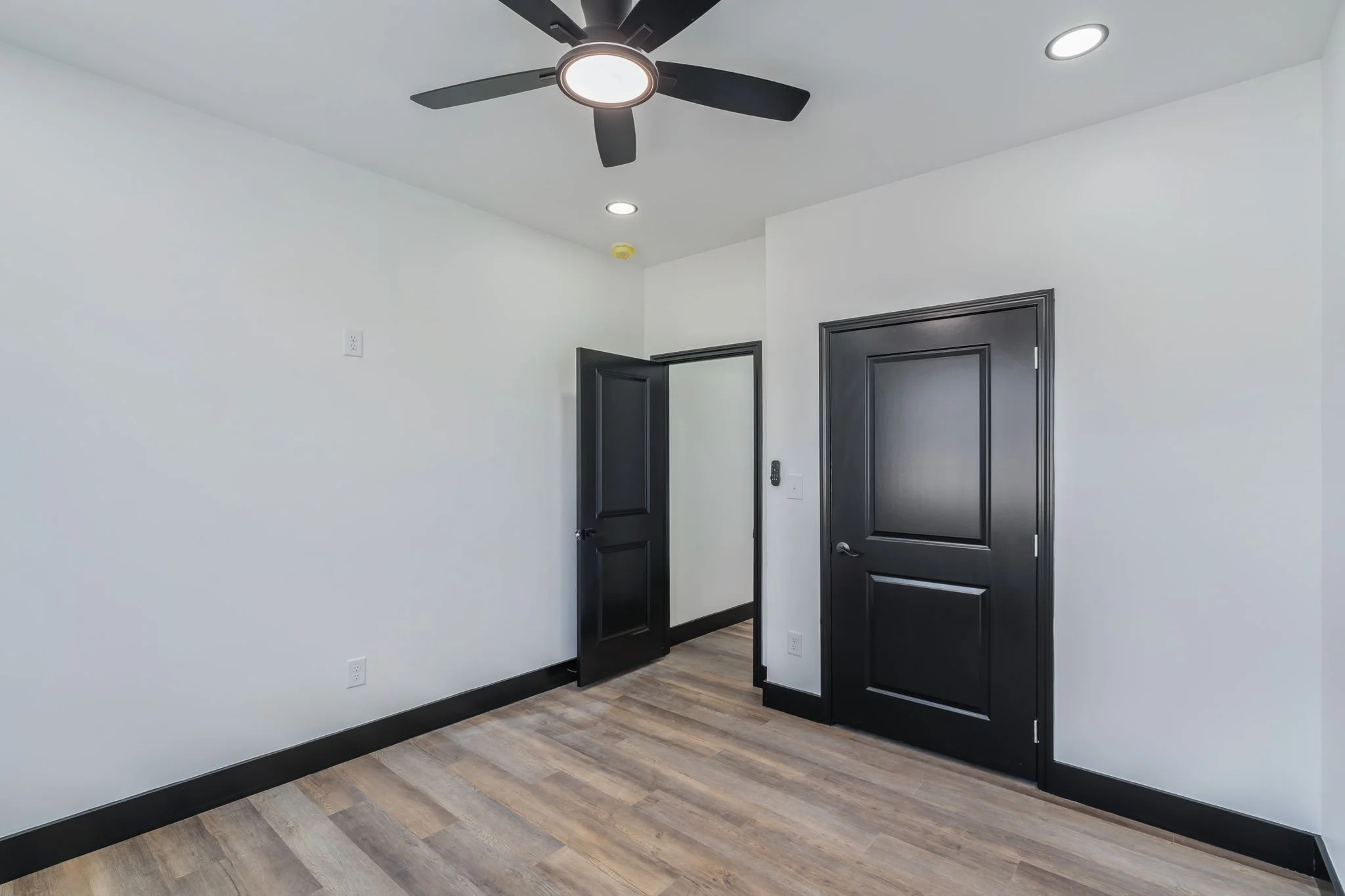 Empty room with white walls, black doors, wood floors, ceiling fan, and recessed lighting.