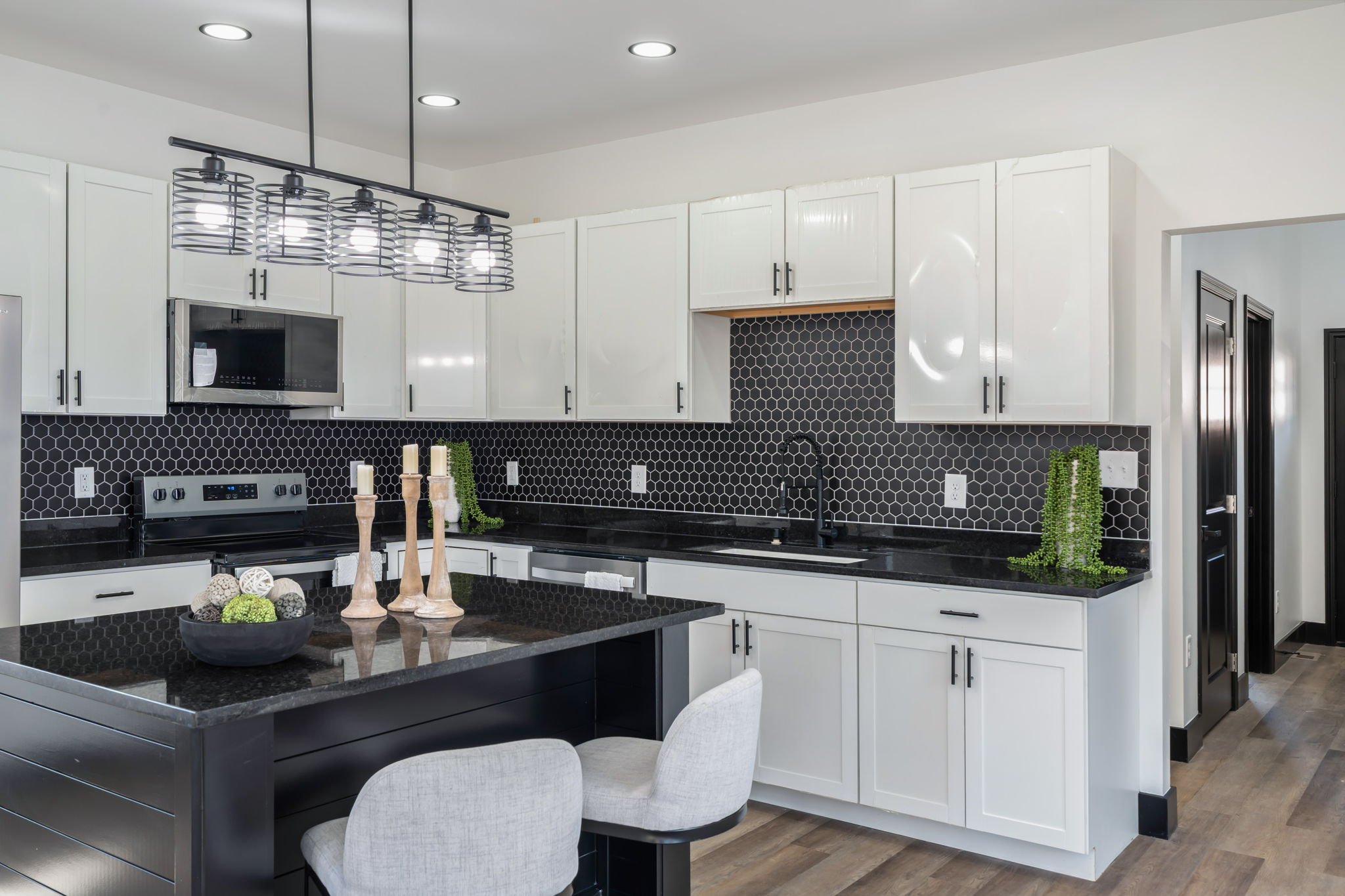 Modern kitchen with white cabinets, black countertops, black hexagonal tile backsplash, and island with light-colored chairs. Decor includes candles, a bowl of balls, and green plants. Overhead lighting fixtures are visible.