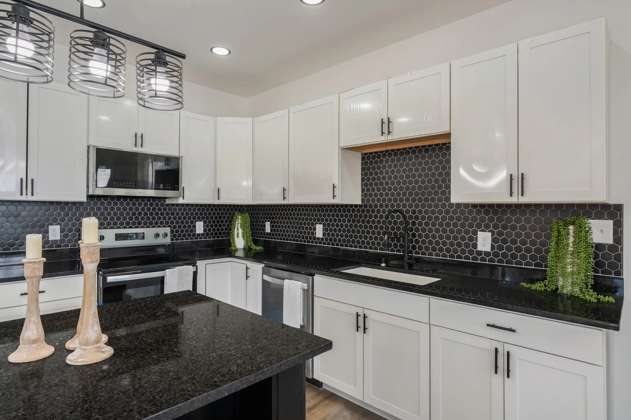 Modern kitchen with white cabinets, black countertops, and a black hexagonal tile backsplash. There are three beige candles on the countertop, plants on the counters, and a black sink with black faucet. A microwave is built into the cabinets, and the