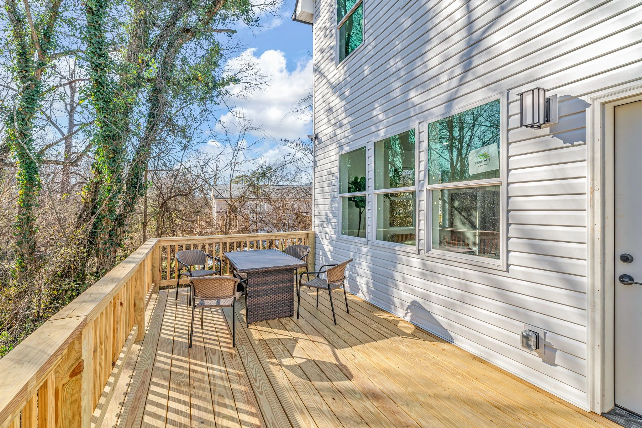 A spacious wooden deck attached to a house with white siding, featuring an outdoor table and four chairs, surrounded by trees with bare branches and a partly cloudy sky.