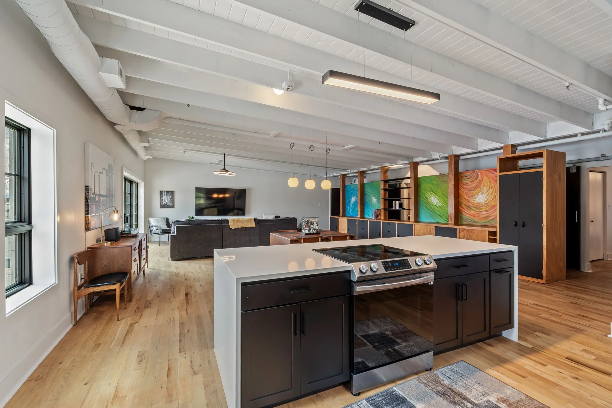 Open-concept living space with a kitchen island, black cabinets, and a living room with a large TV and gray sofa, featuring white-painted ceiling beams and wood flooring.