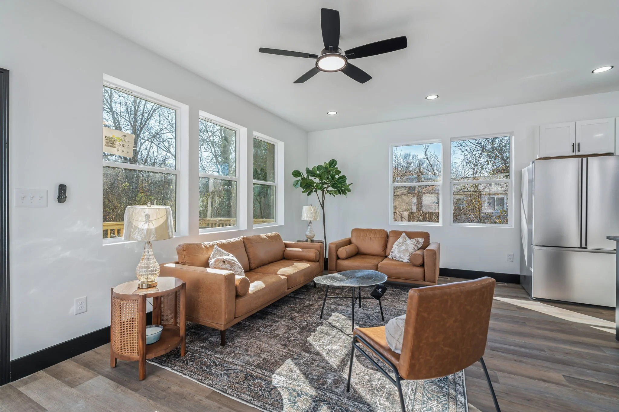 Bright living room with white walls and large windows, featuring brown leather sofas, a patterned area rug, a small round coffee table, a side table with a lamps, a potted plant, and a black ceiling fan.