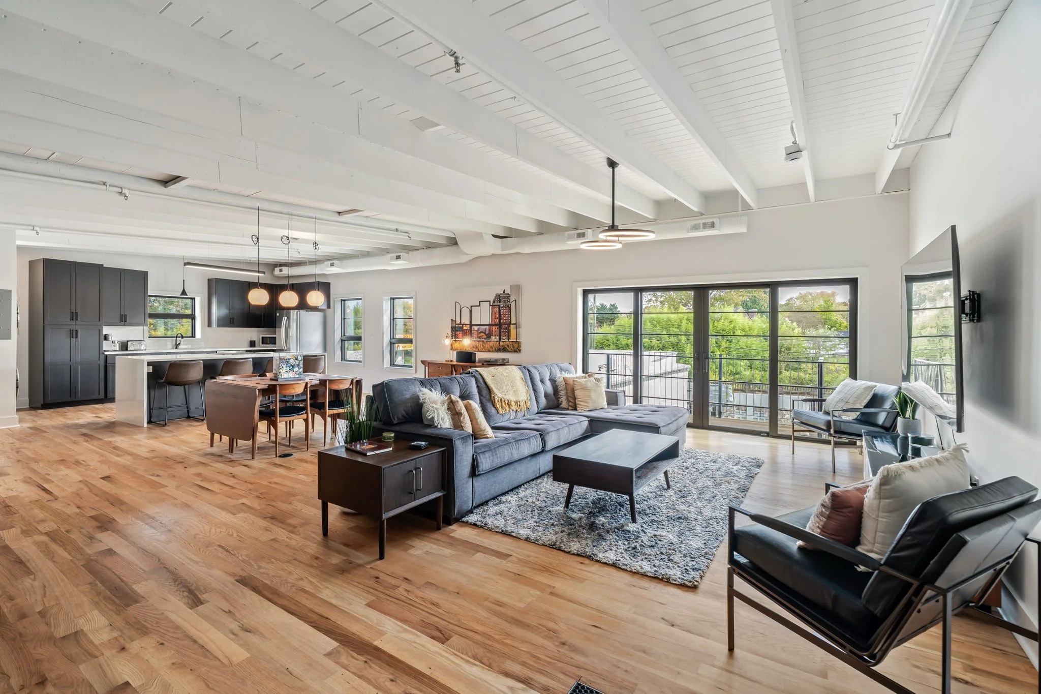 Open-concept living room and kitchen with hardwood floors, large windows, gray sofa, black armchair, and dining table with chairs.