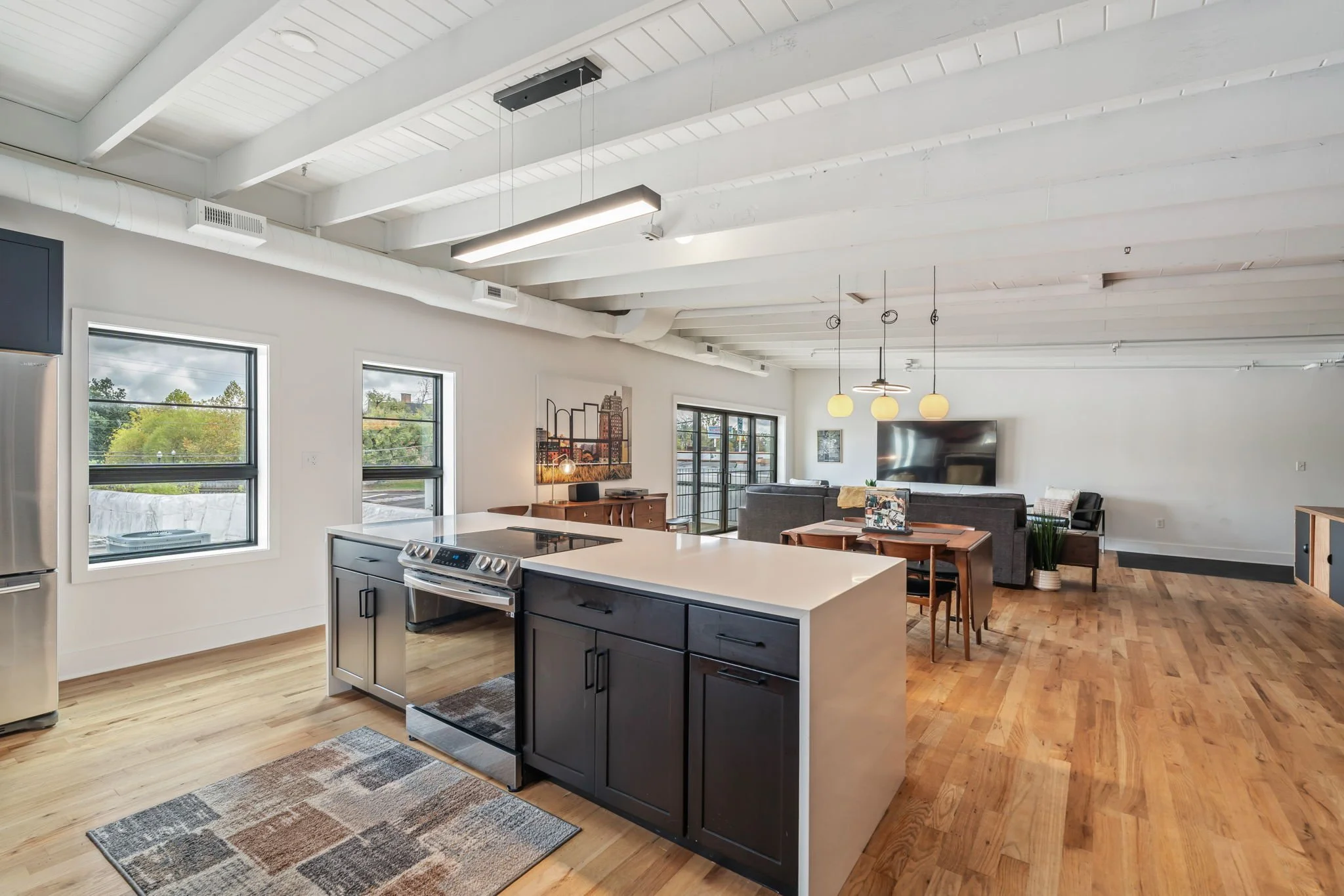Open-concept living space with a kitchen, dining, and living area, featuring white walls, large windows, hardwood floors, and modern furniture.
