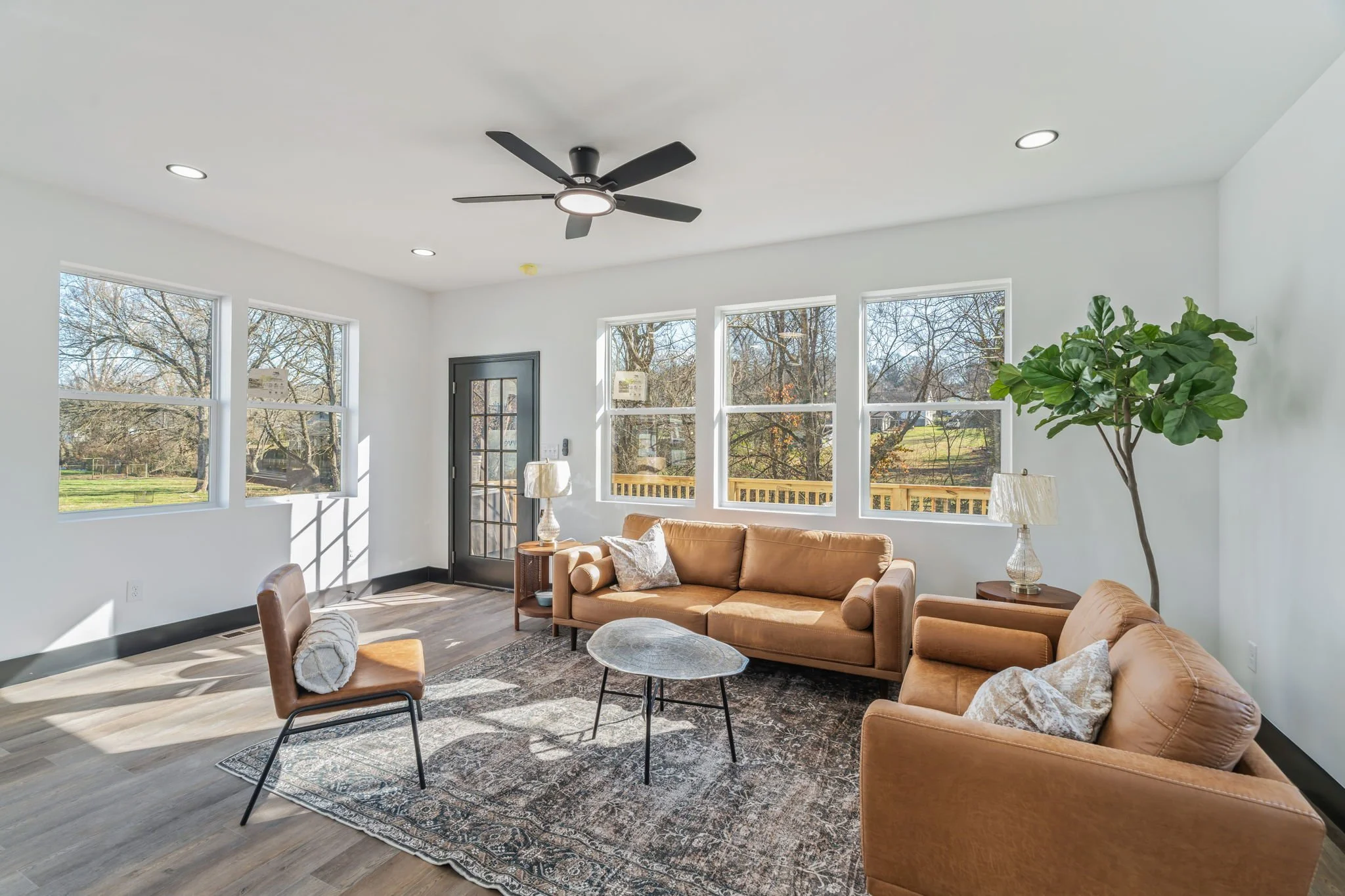 Bright living room with beige sofas, a wooden coffee table, a large green potted plant, three large windows, and a ceiling fan.