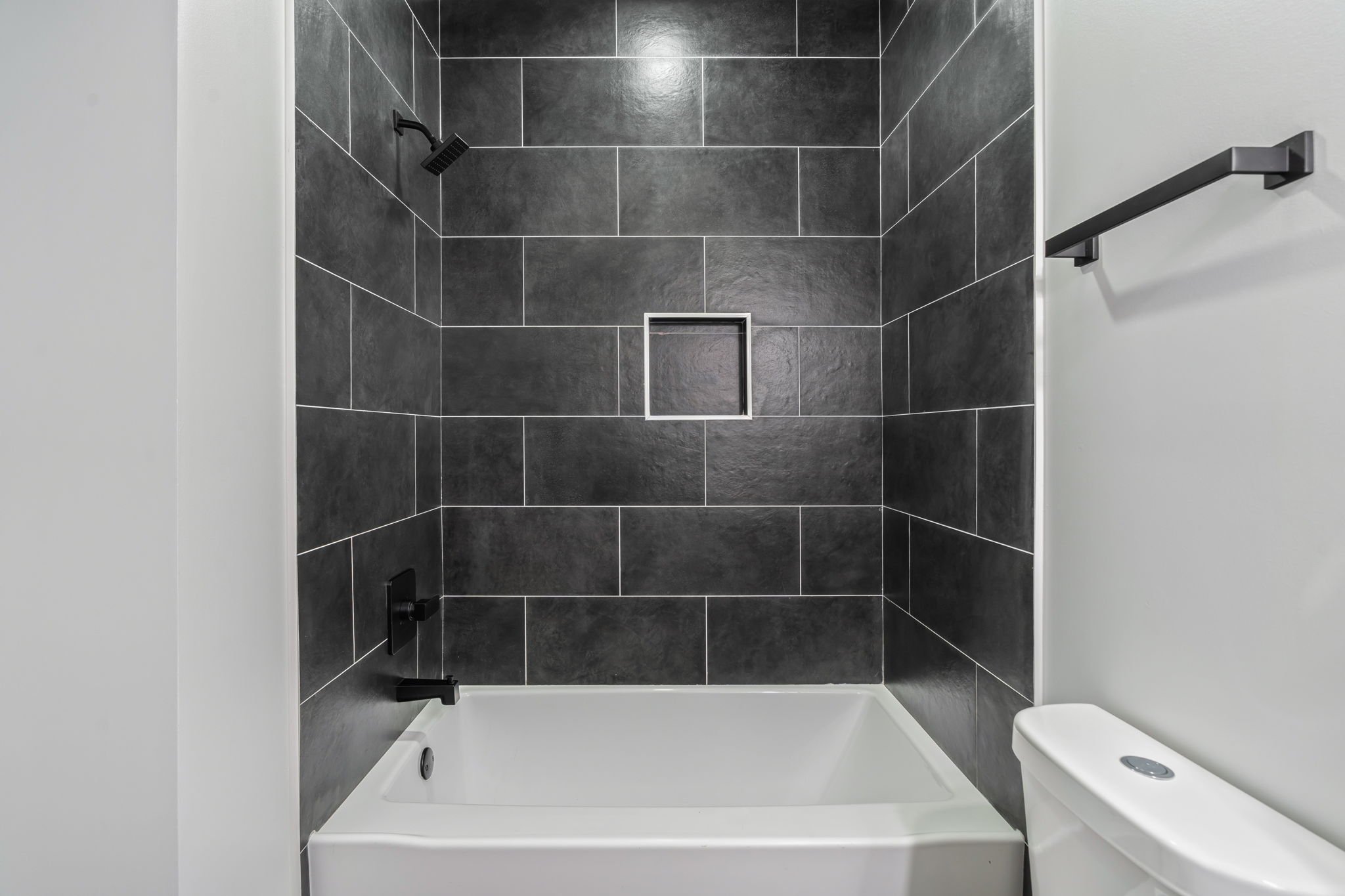 Bathroom shower with black tile walls, a built-in niche, a black showerhead, a black faucet, a towel bar on the white wall, and a white bathtub with a toilet beside it.