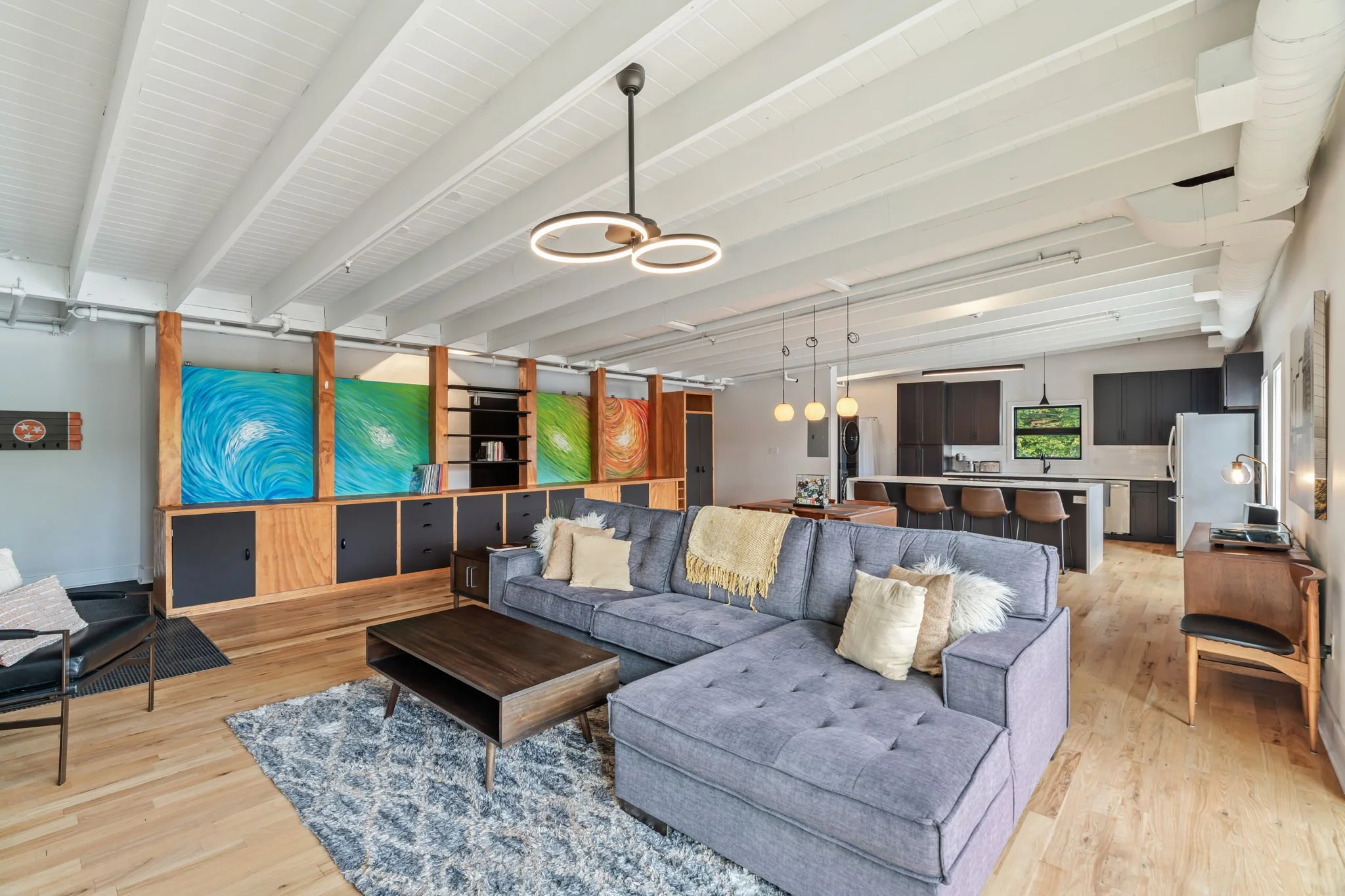 Open-concept living room with gray sectional sofa, wooden coffee table, colorful abstract paintings on wood-paneled wall, and modern kitchen in background with black cabinets and bar stools.