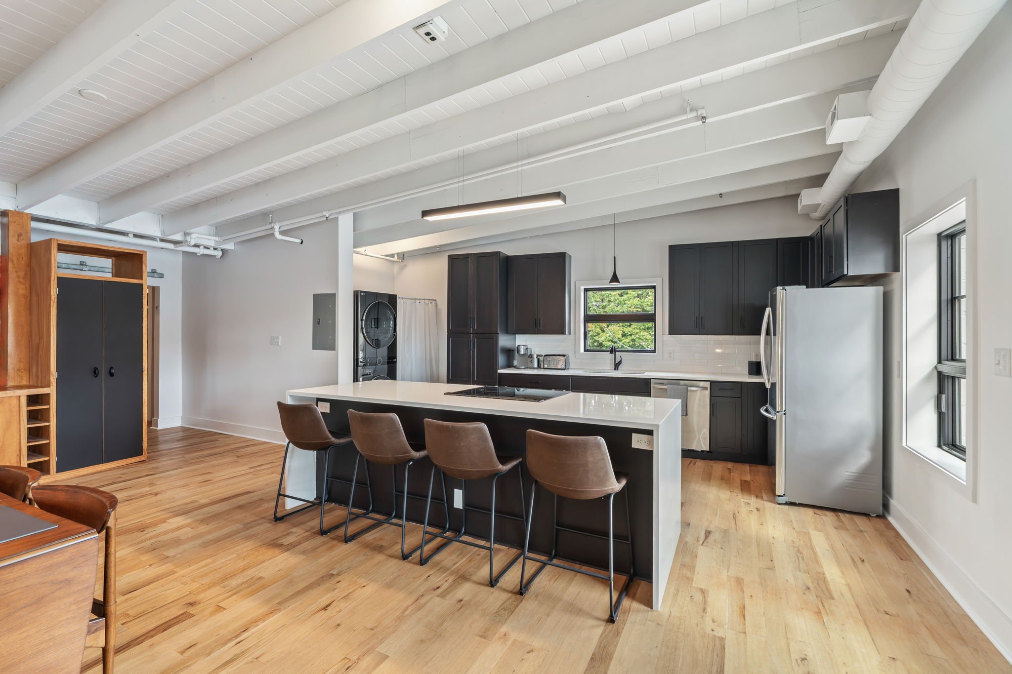 Modern kitchen with black cabinets, white countertops, a large island with four brown chairs, stainless steel appliances, wood flooring, and windows allowing natural light.