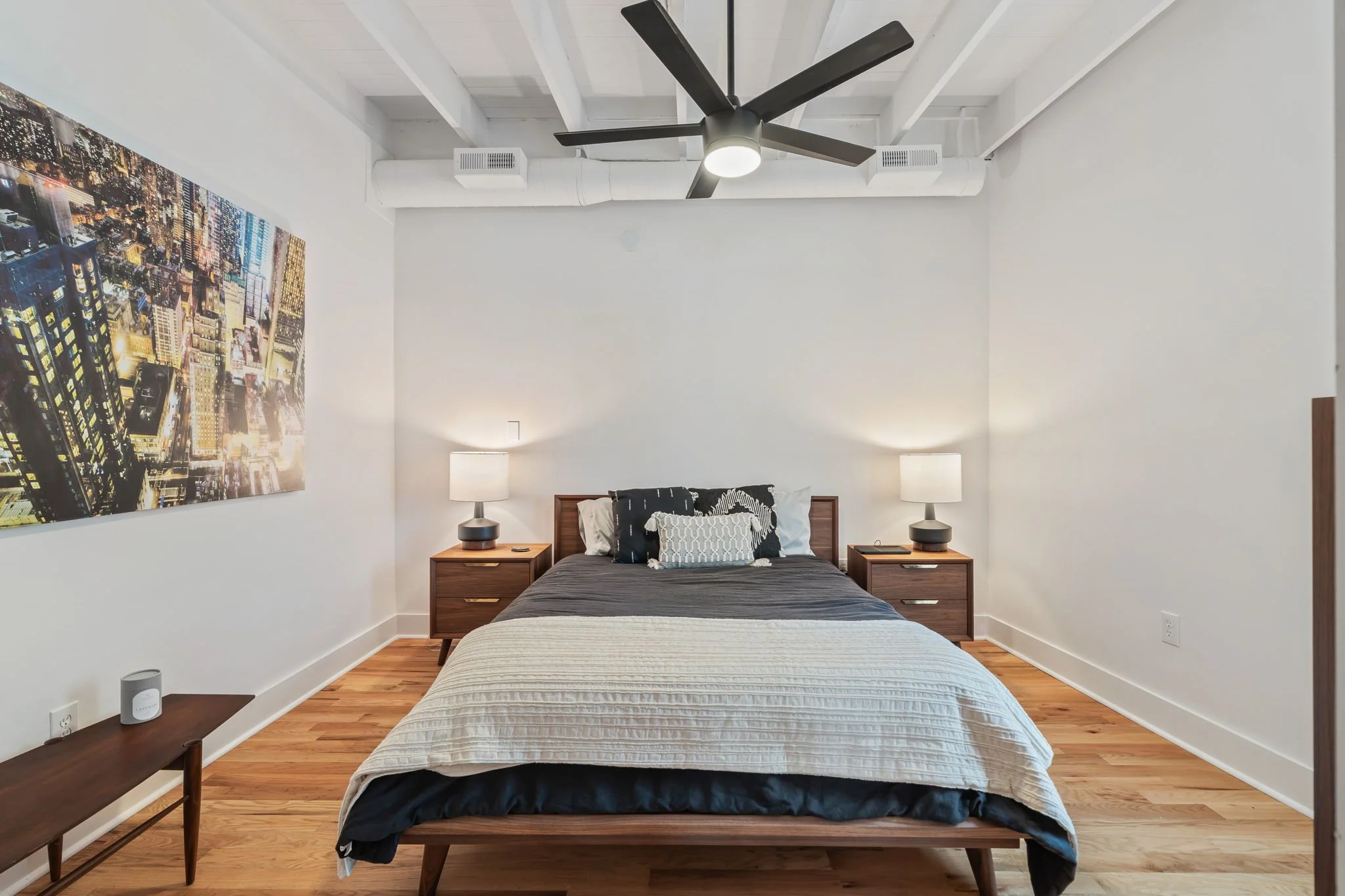 A bedroom with a bed, two matching nightstands with lamps, a large cityscape art piece on the wall, hardwood flooring, and a ceiling fan with lights.